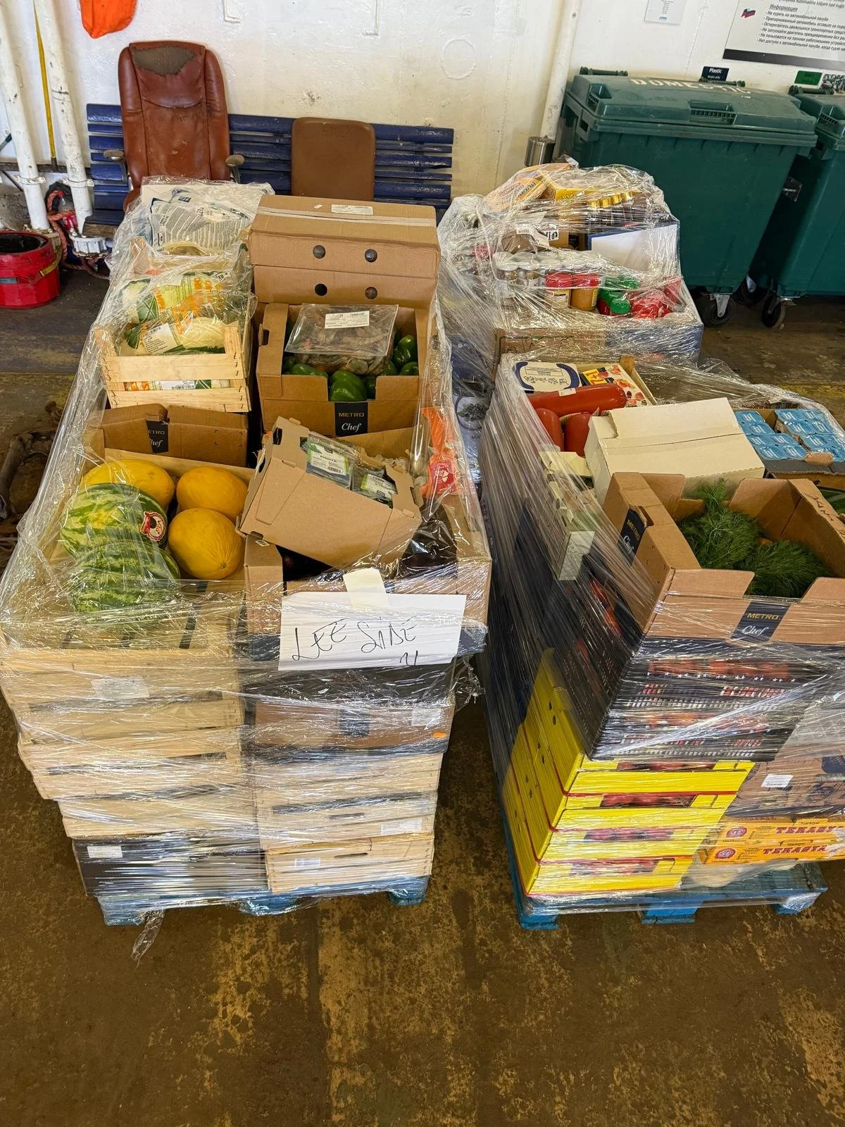 Stacks of boxed produce and food items wrapped in plastic wrap, including melons, green peppers, and cherry tomatoes, in a warehouse or storage area.