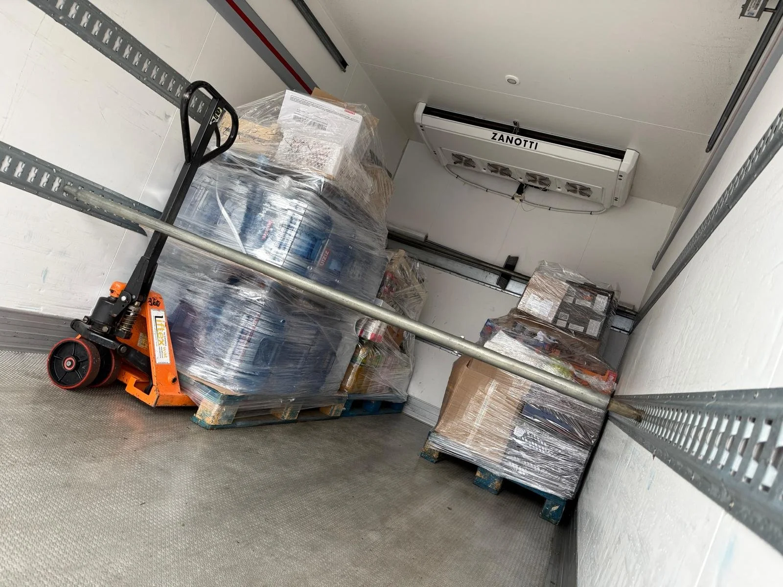 The inside of a storage unit with pallets of water bottles and boxes wrapped in plastic, a hand truck, and an air conditioning vent on the ceiling.