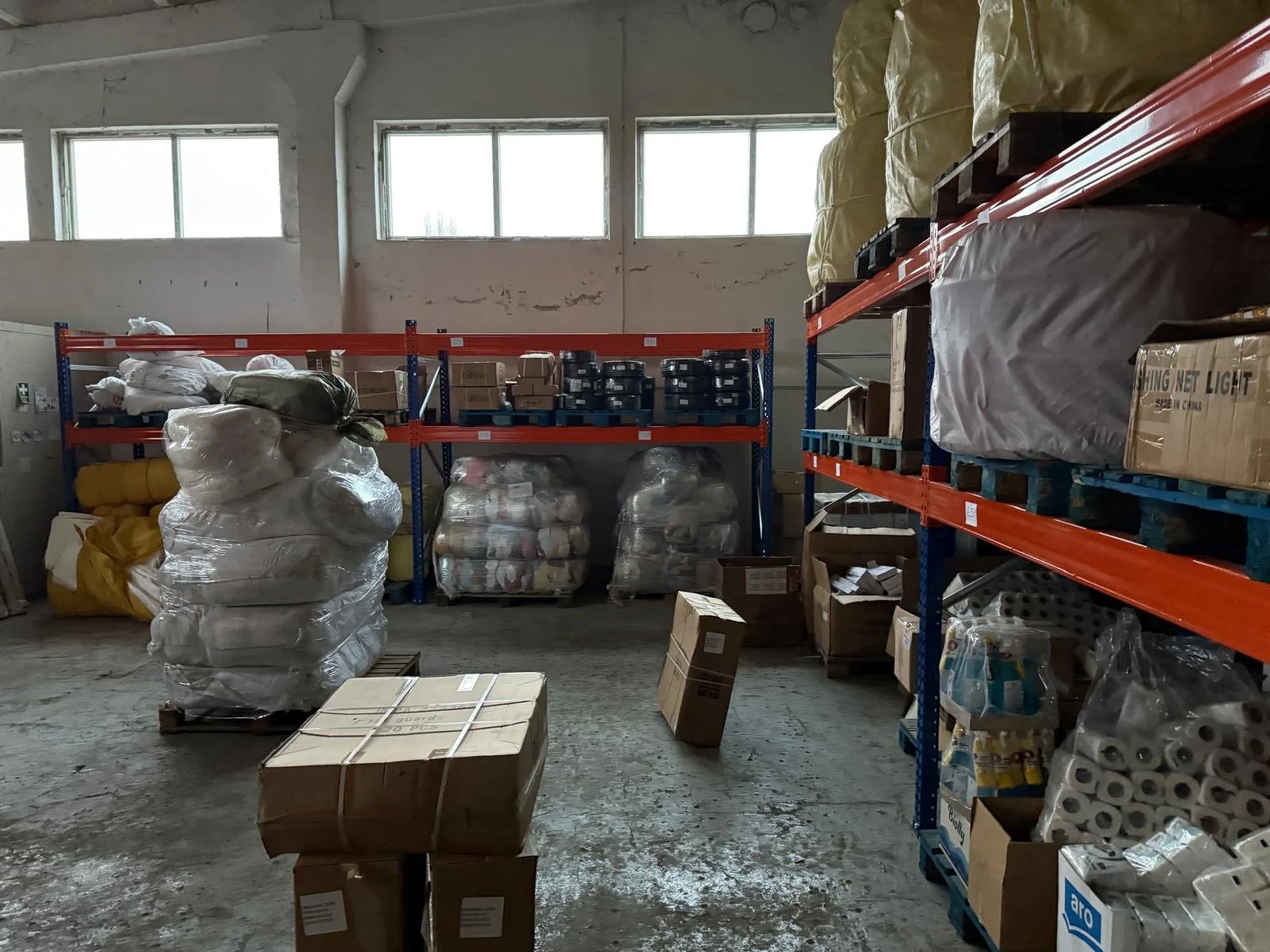 Interior of a storage room with metal shelving units holding boxes, bags, and supplies like rolls of paper towels. The room has high windows and a concrete floor.