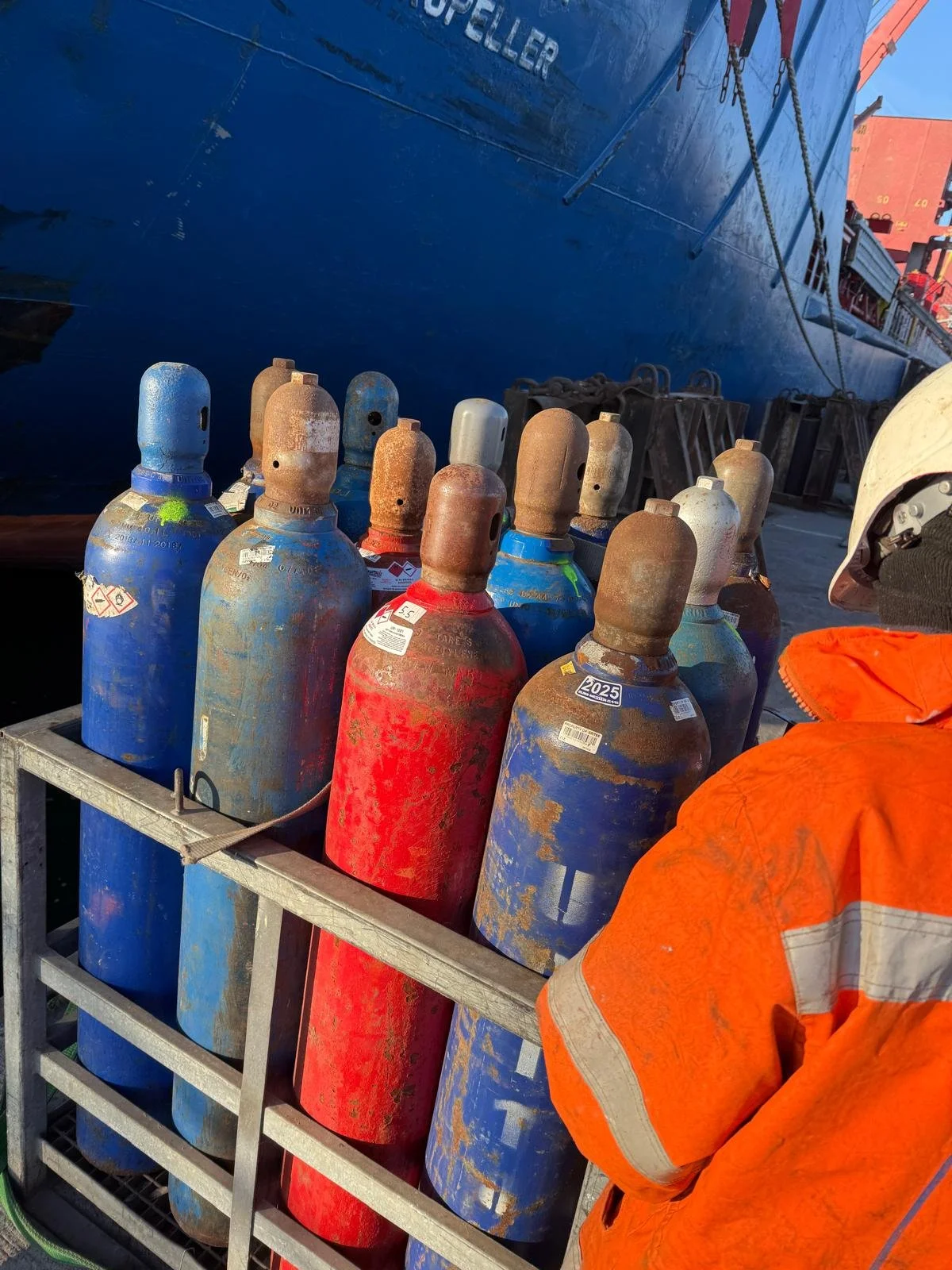 Several weathered oxygen tanks in red, blue, and white colors mounted on a metal cart, with a person in an orange safety jacket and helmet nearby, and a large ship in the background at a dock.