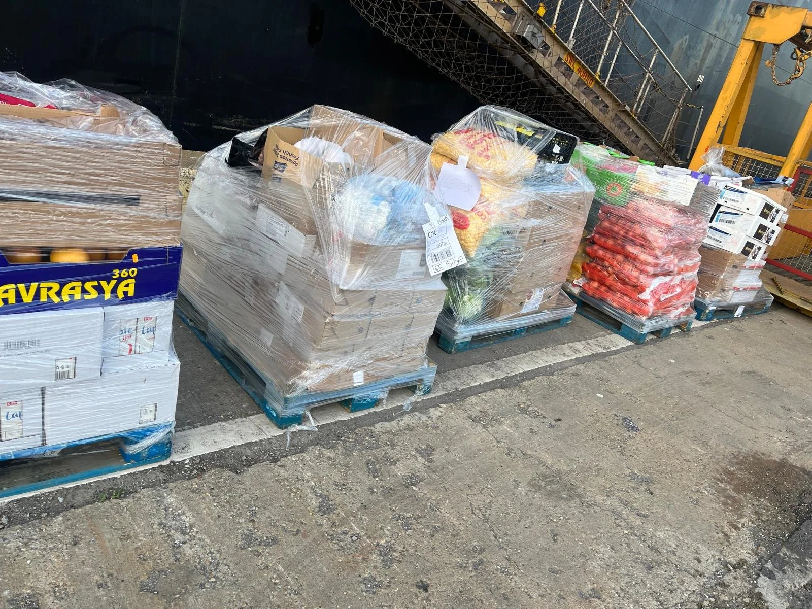 Pallets of packaged goods including boxes of eggs, crushed packets of food, and other supplies on a loading dock with a black container and yellow crane in the background.