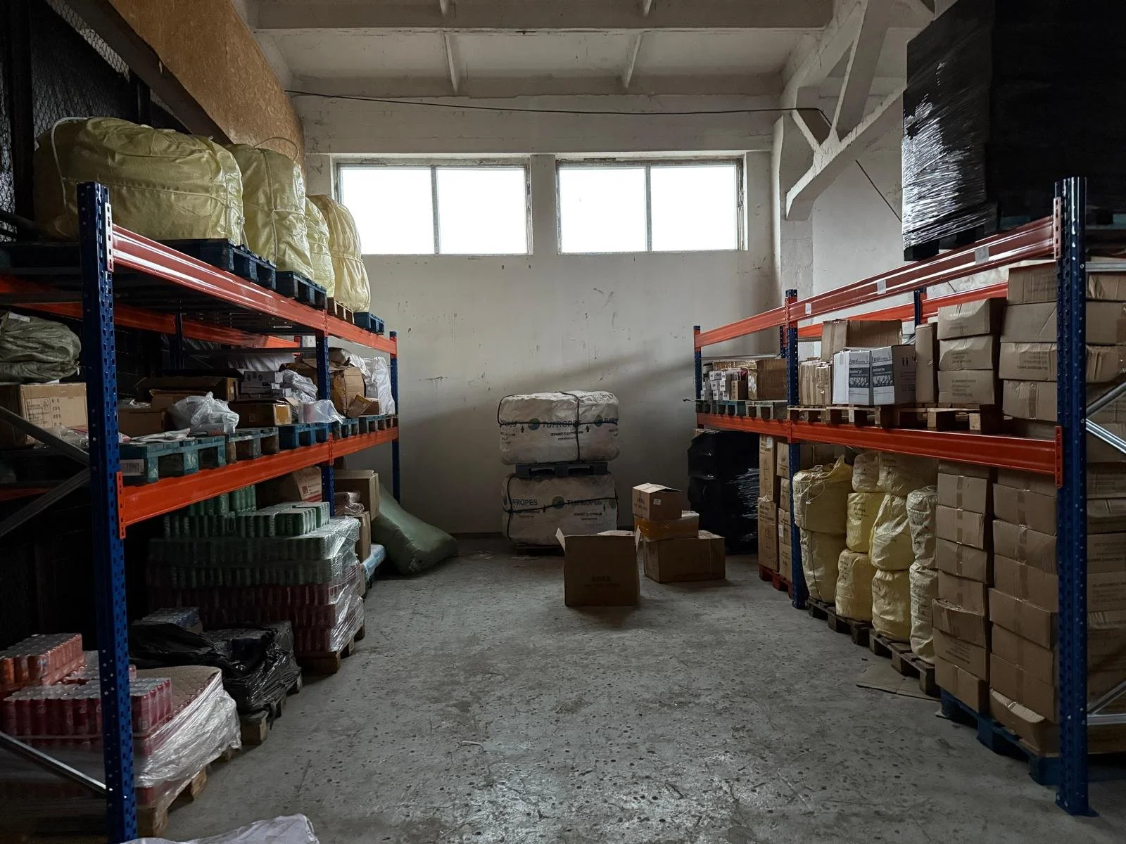 Storage room with metal shelves filled with various packages, boxes, and bags, some wrapped in plastic, under a high ceiling with windows near the top letting in natural light.