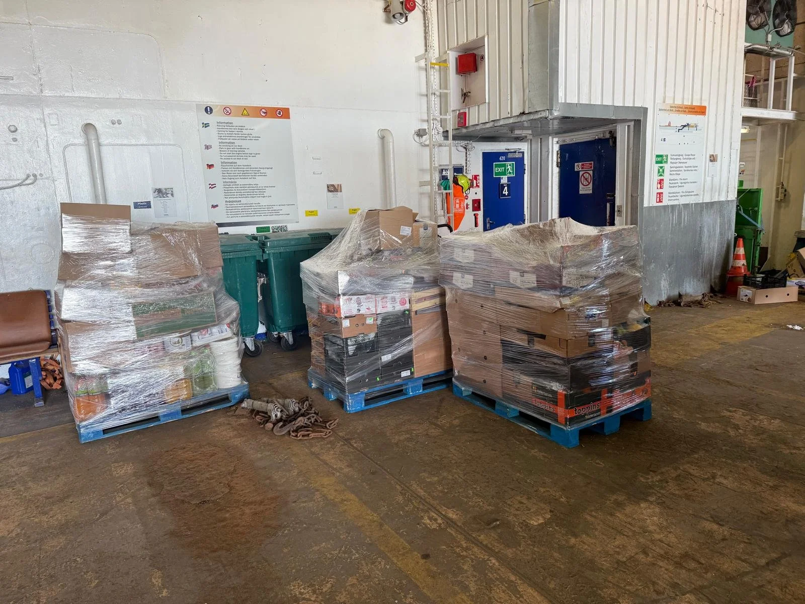 Pallets of boxed goods and supplies wrapped in plastic are stacked on a warehouse floor near a wall with safety signs and a large industrial structure, with trash and equipment in the background.