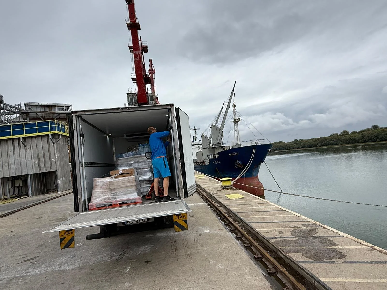 A man in blue clothes loading pallets from a truck onto a dock near a river, with a blue and white ship moored nearby and a large red crane in the background.