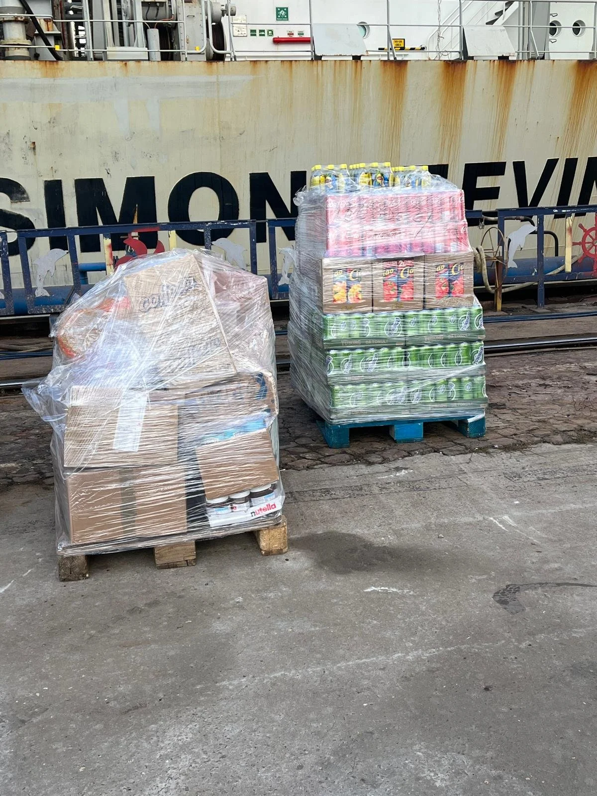 Two pallets of bottled beverages and canned goods wrapped in plastic on concrete ground near a ship with large writing, against a rusty metal railing.