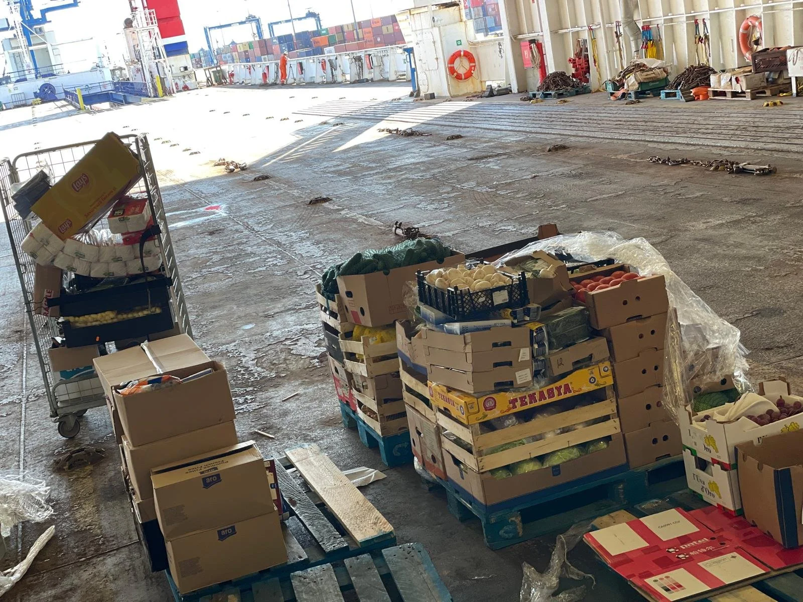 Pallets of various produce and boxed goods on the deck of a cargo ship at port, with shipping containers and industrial equipment visible in the background.