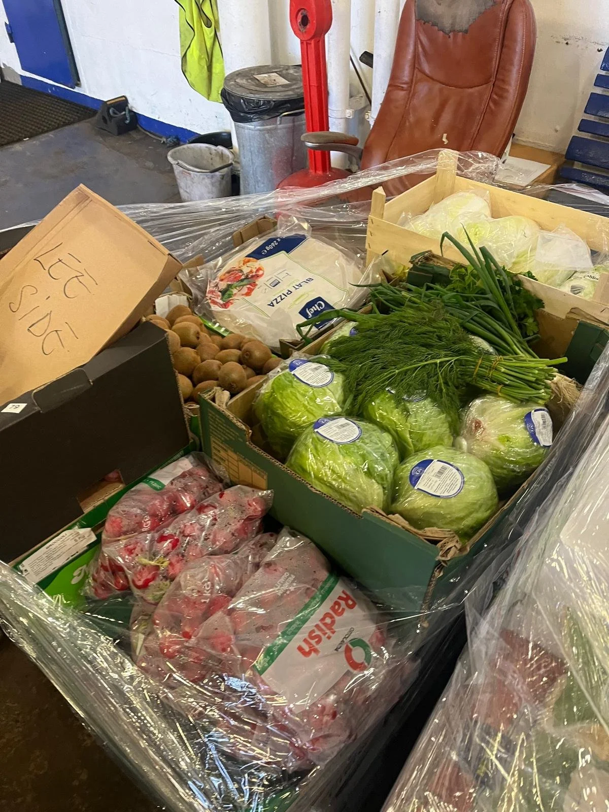 A box of fresh vegetables including lettuce, green onions, dill, potatoes, and strawberries on a table in a warehouse or produce area.