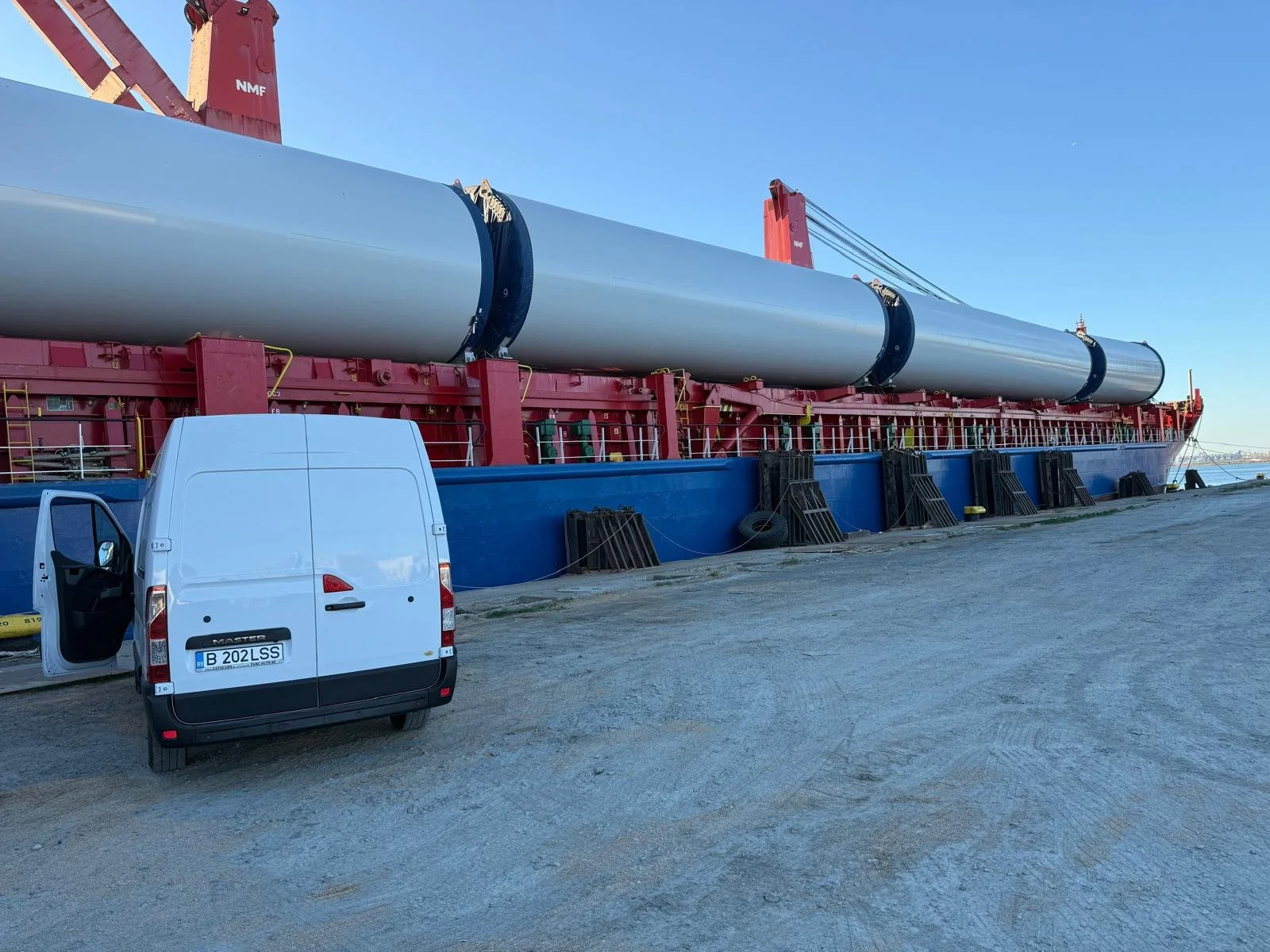 Large offshore wind turbine blade on a barge with a white van nearby, set against a clear blue sky.