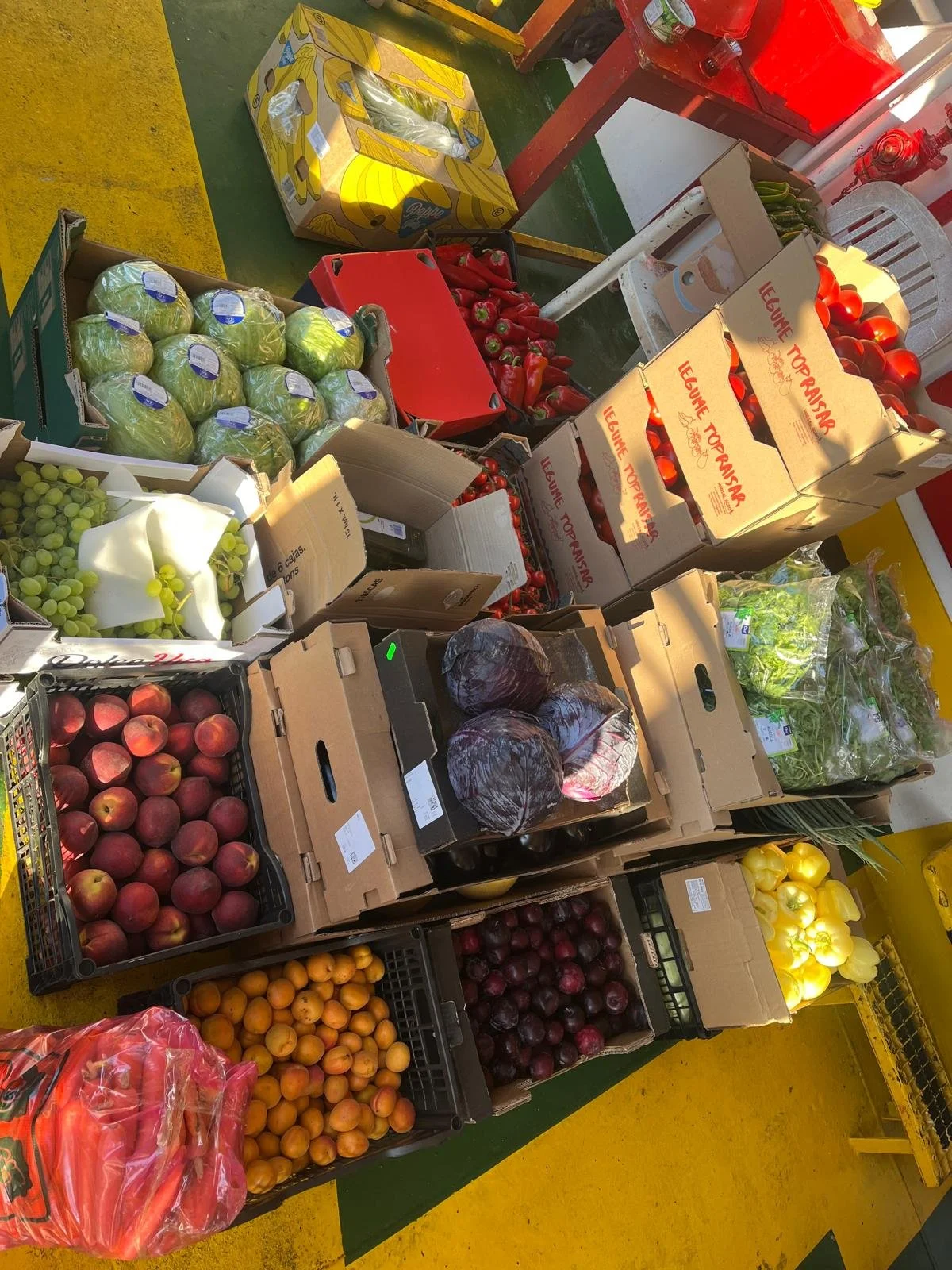 A variety of fresh fruits and vegetables on display at a market stand, including apples, oranges, tomatoes, cabbage, and peppers, arranged in crates and boxes.