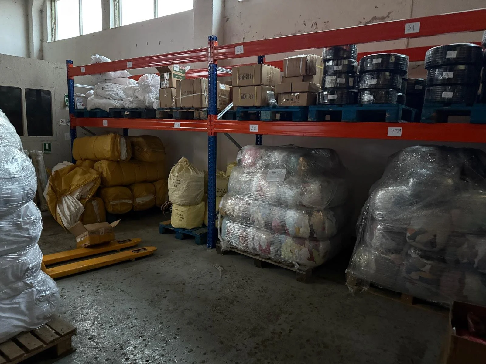 Shelves in a warehouse with boxes, bags, and palletized materials, some covered in plastic wrap.