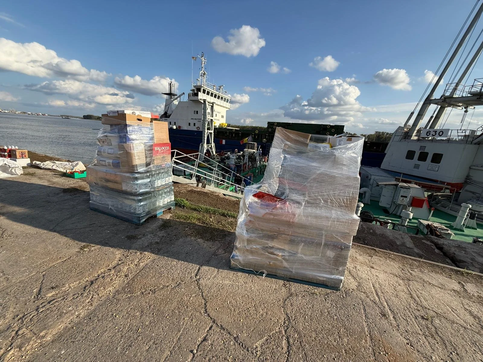Stacks of wrapped cardboard boxes and supplies on the dock near boats and ships at a harbor.