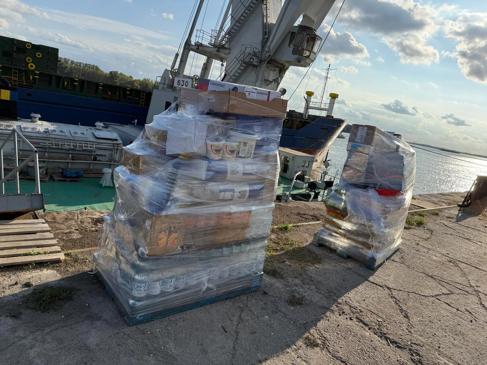 Pallets of boxed and canned supplies wrapped in plastic at a dock near the water, with boats and a crane in the background.
