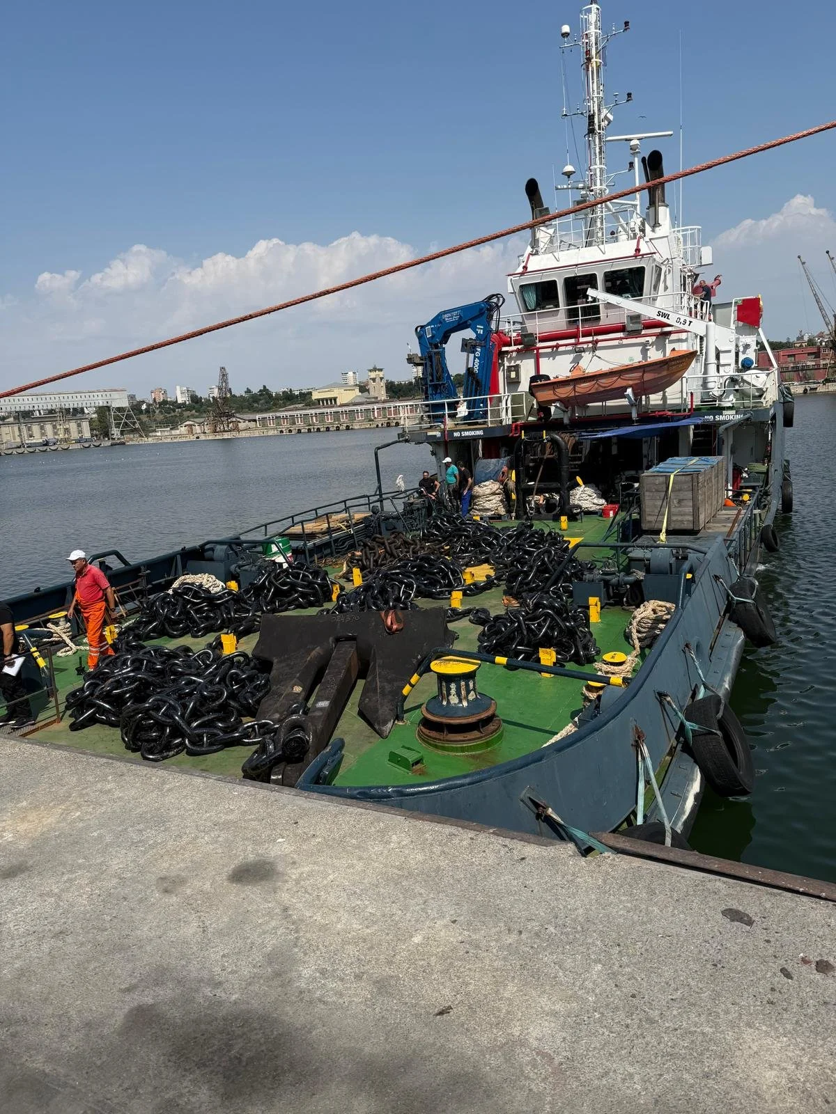 A large boat docked at a port with big chains and equipment on its deck. Several workers are handling the equipment. The boat has a white upper structure with various antennas and a small orange boat on its side.