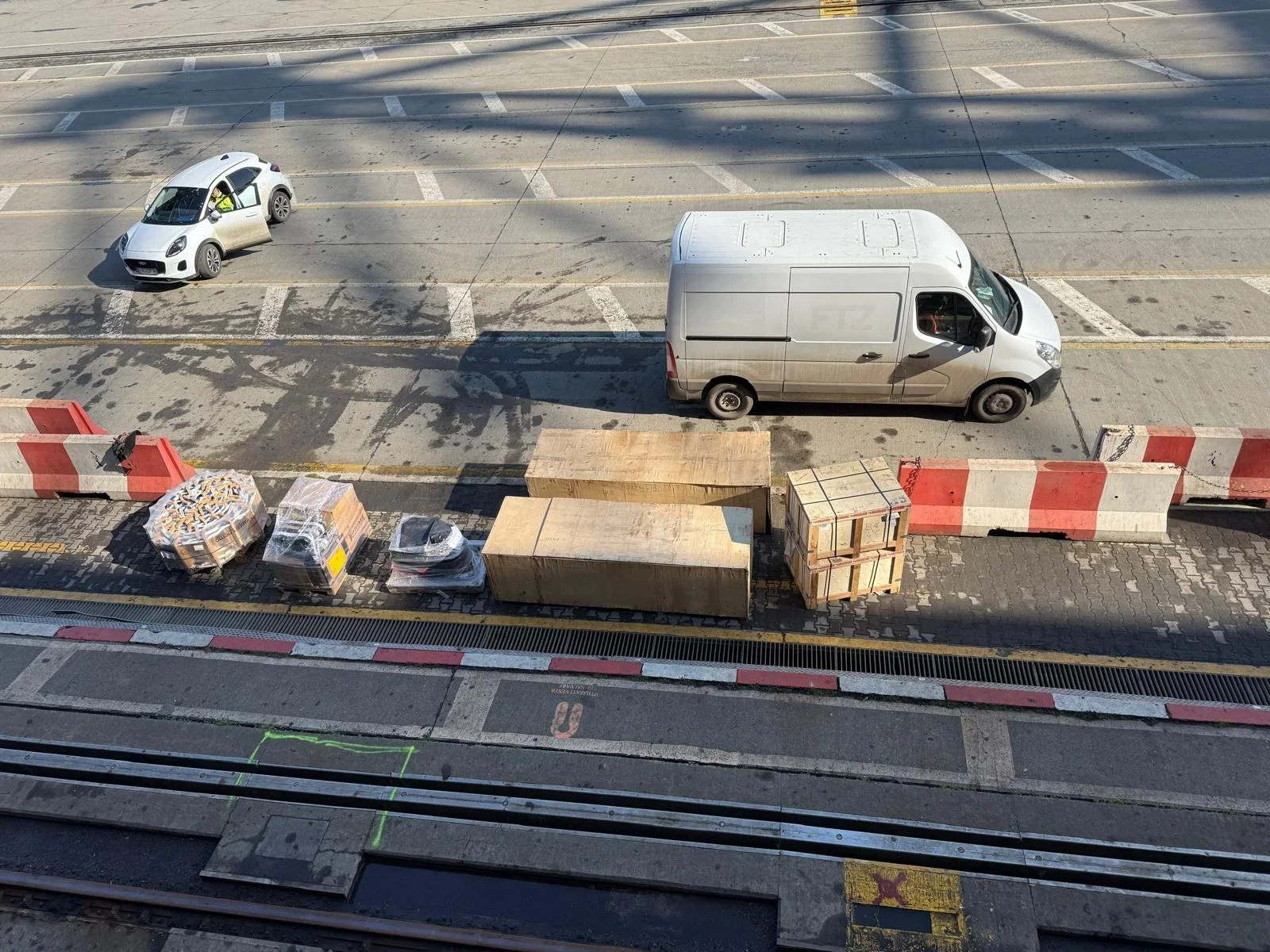 A street view showing a parked gray delivery van, a white compact car with an open door, construction materials, and barriers along the sidewalk under shadows.