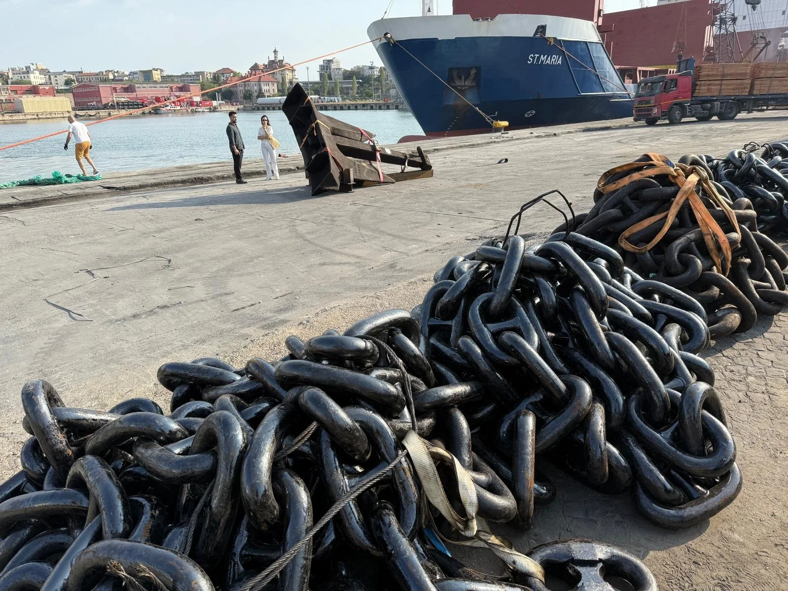 Large black metal chains on dock near ships with people walking by and buildings across the water.