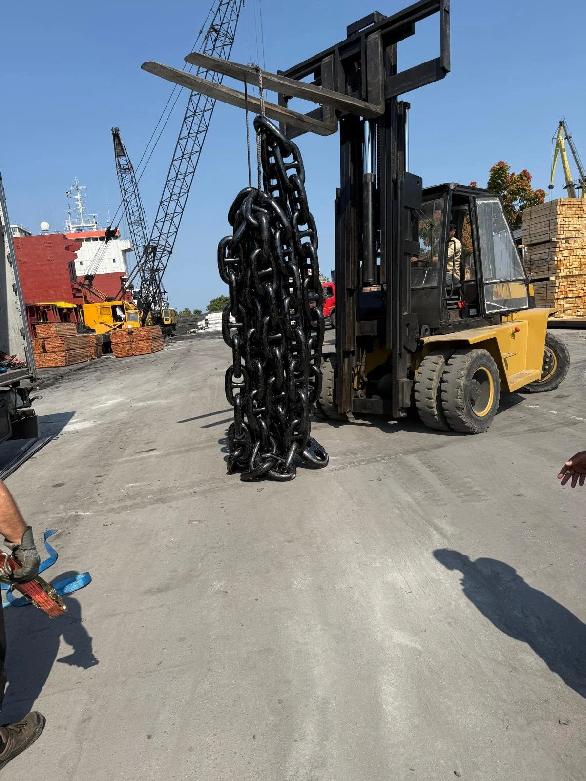 A forklift lifting a large, thick chain at an industrial port with ships, cranes, and stacks of wood in the background.