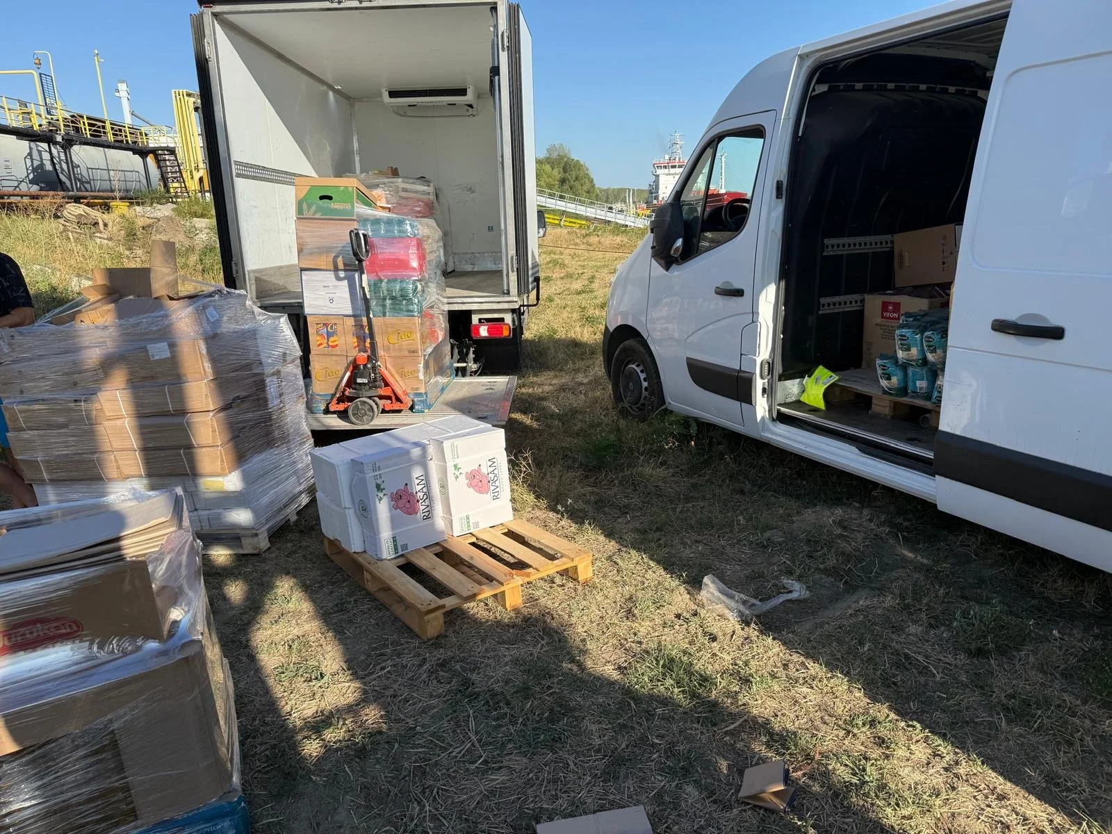 Two trucks, one with an open side door revealing boxes and supplies inside, parked on grass near industrial equipment, with pallets of goods and cardboard boxes on the ground outside.