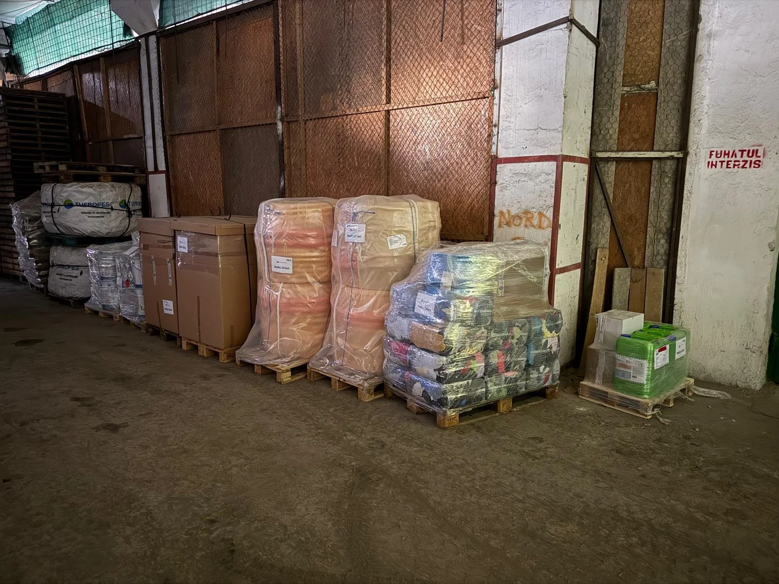 Pallets of packaged goods, including boxes and bags, stored in an industrial warehouse with a dirt floor and metal fencing.