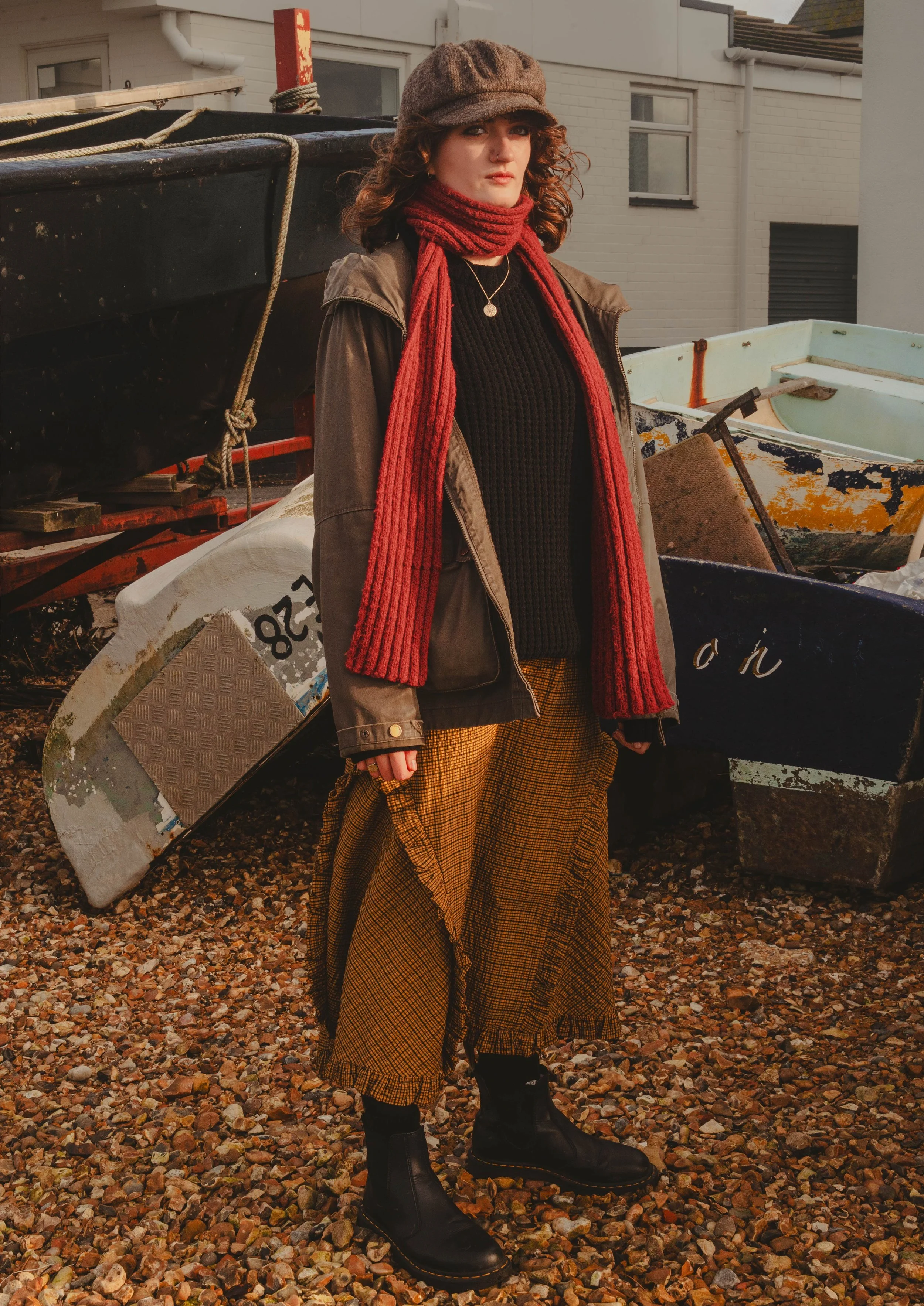 A woman with curly hair and a brown hat outdoors. She wears a red scarf, black sweater, brown jacket, yellow plaid wide-leg pants, and black boots. She stands on a gravel surface with boats and a building in the background.