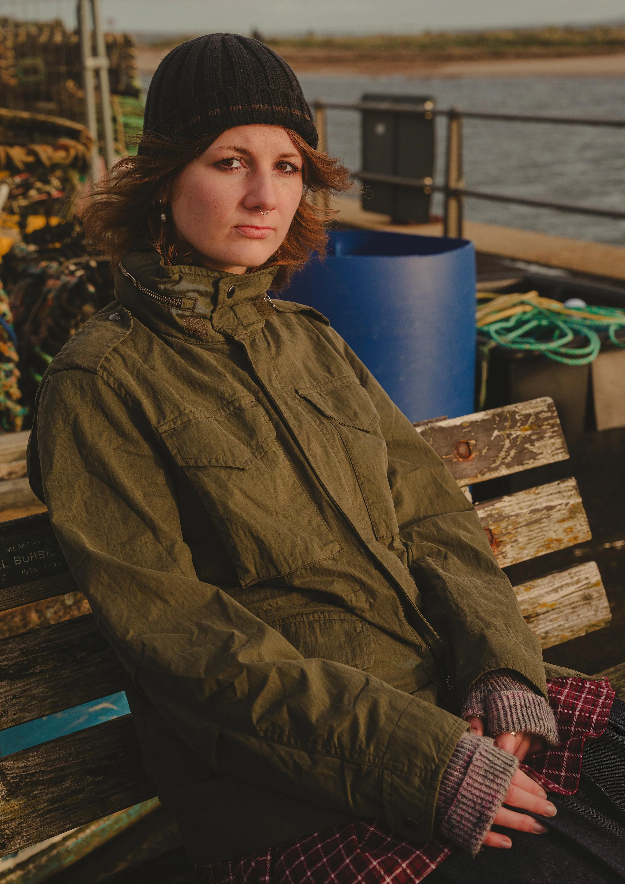 A young woman with brown hair wearing a black knit hat, green jacket, and plaid skirt, sitting on a wooden bench by a waterfront with her hands clasped in her lap.