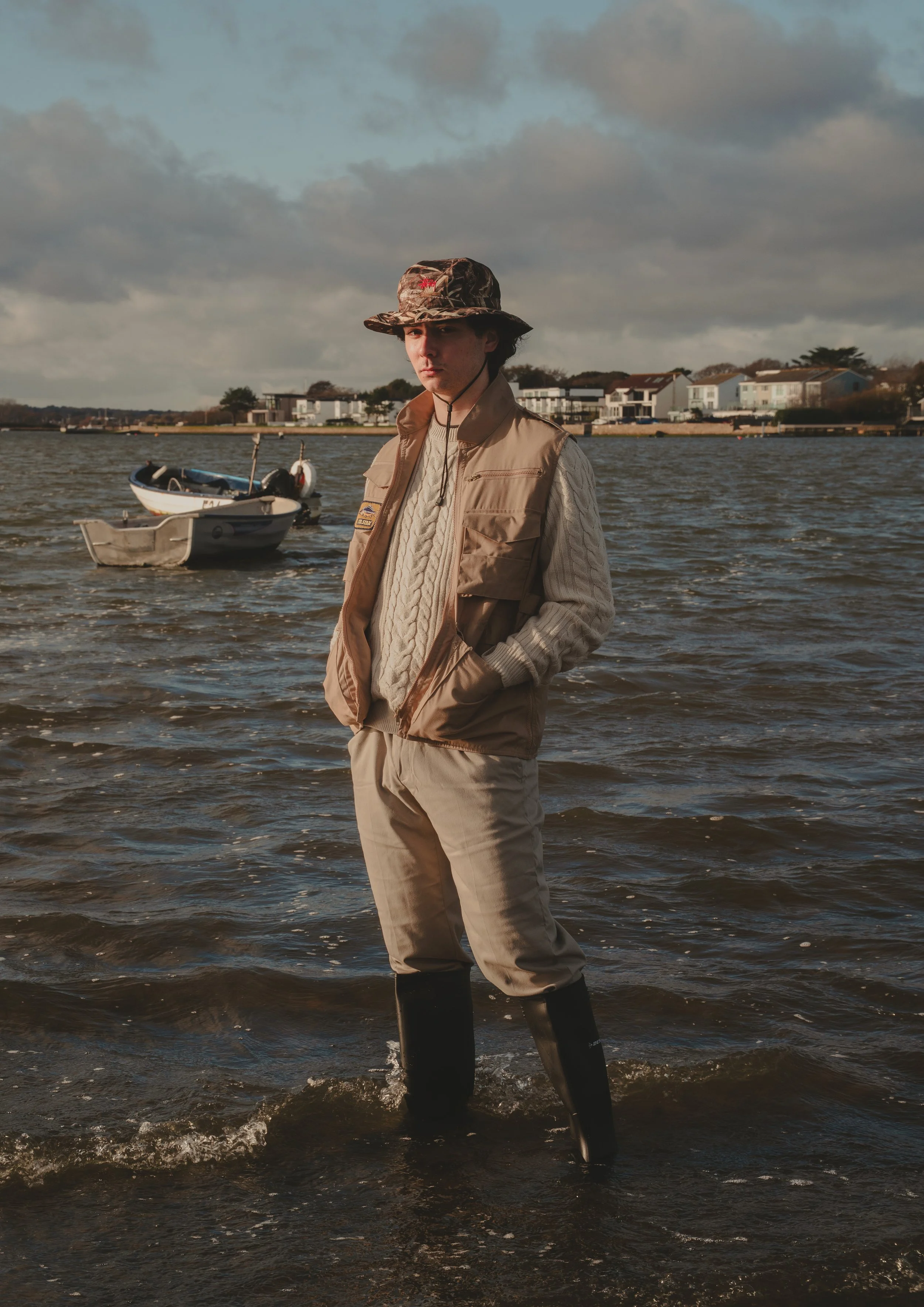 A young man standing in shallow water near the shoreline, wearing a beige vest, a cable-knit sweater, and a camouflage bucket hat, with boats floating on the water and houses visible on the distant shore under cloudy skies in the background.