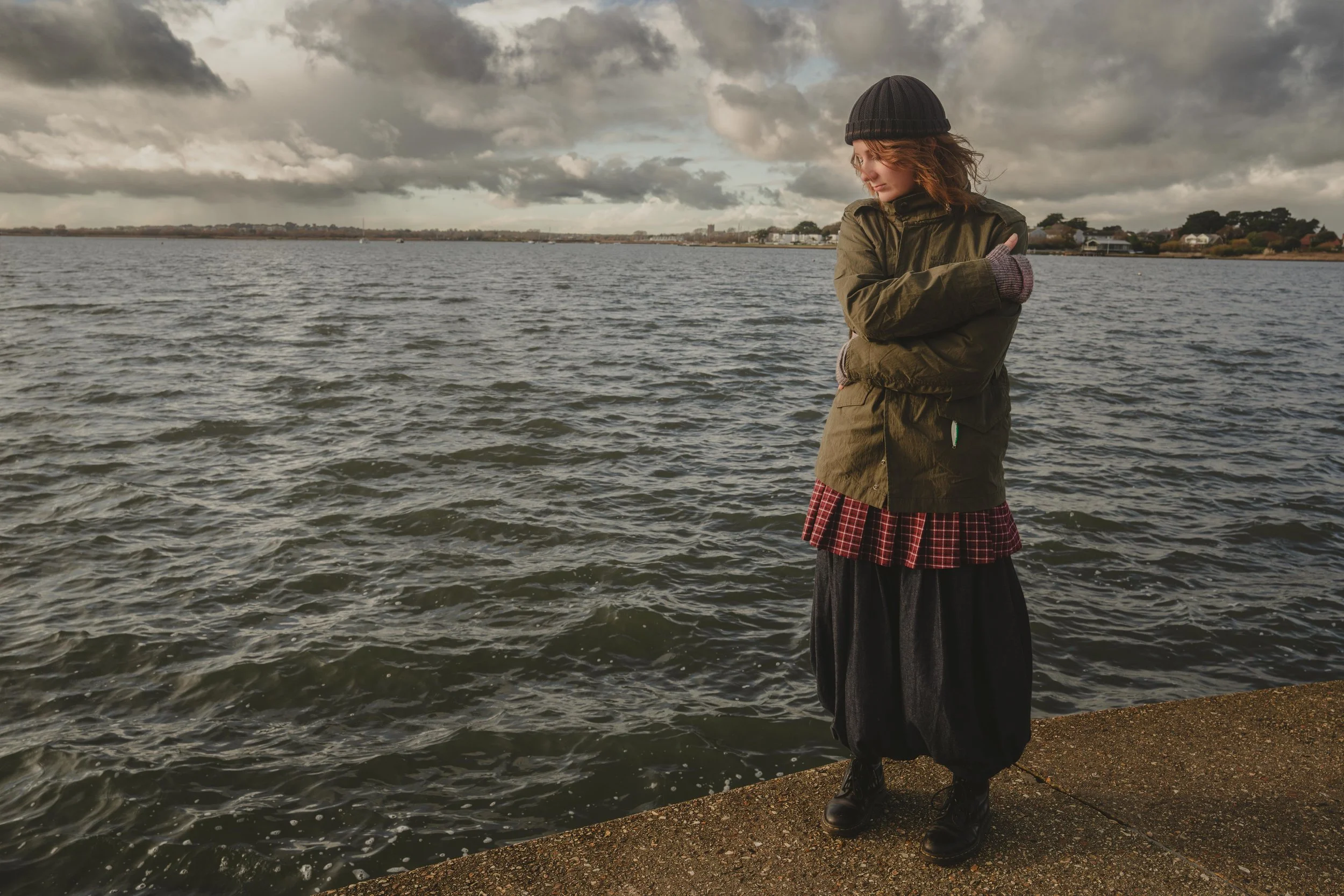A woman stands by a body of water during cloudy weather, wearing a black beanie, green jacket, and a red plaid skirt.