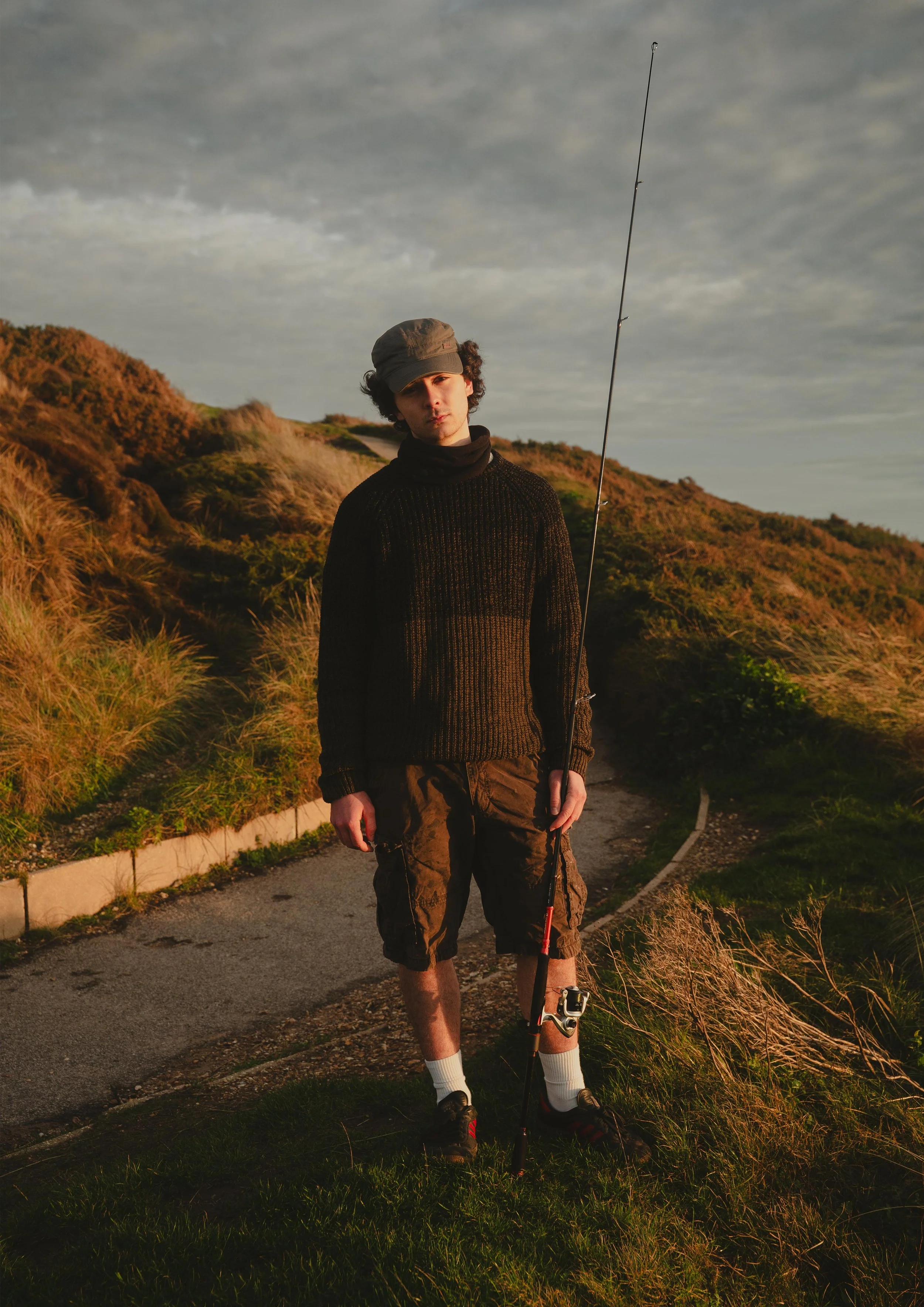 Young man standing on a paved pathway with green grass and bushes, holding a fishing rod, wearing a hat, black sweater, shorts, socks, and sneakers, under a cloudy sky during sunset.