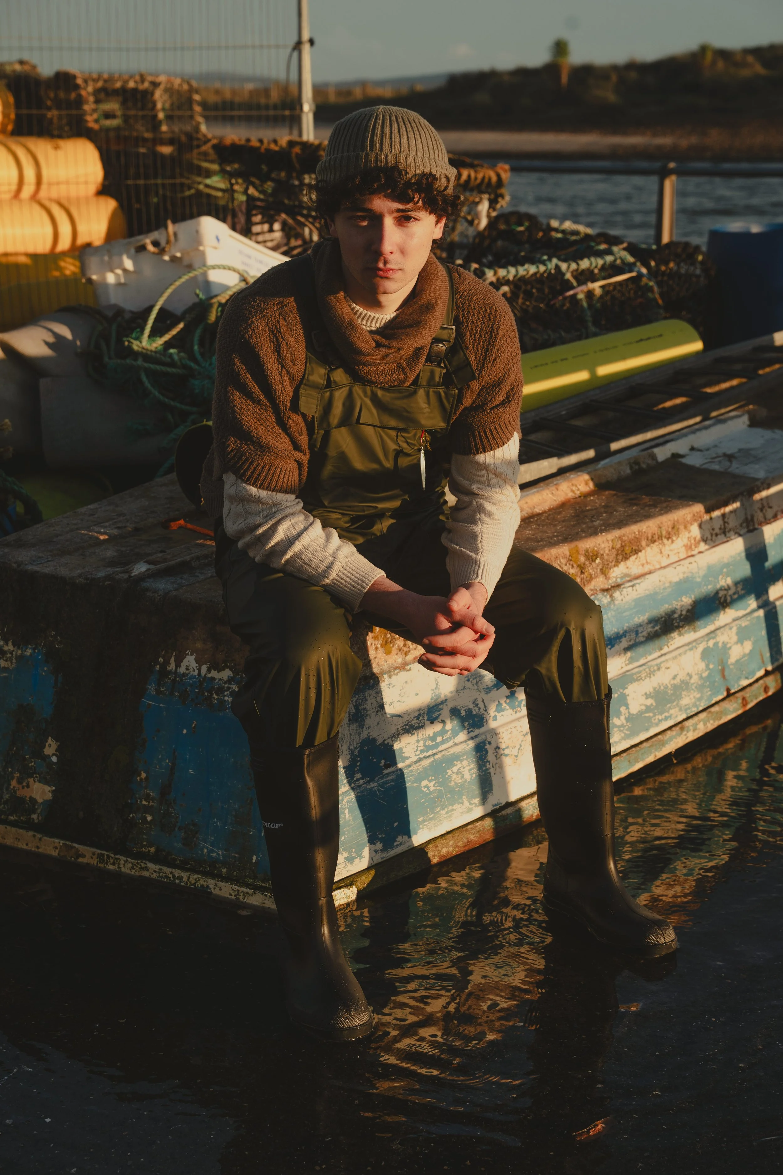 Young man sitting on a weathered boat at the water's edge, dressed in waterproof boots, olive overalls, a knitted hat, and layered clothing, with a backdrop of a river, a fence, and land in the late afternoon sunlight.