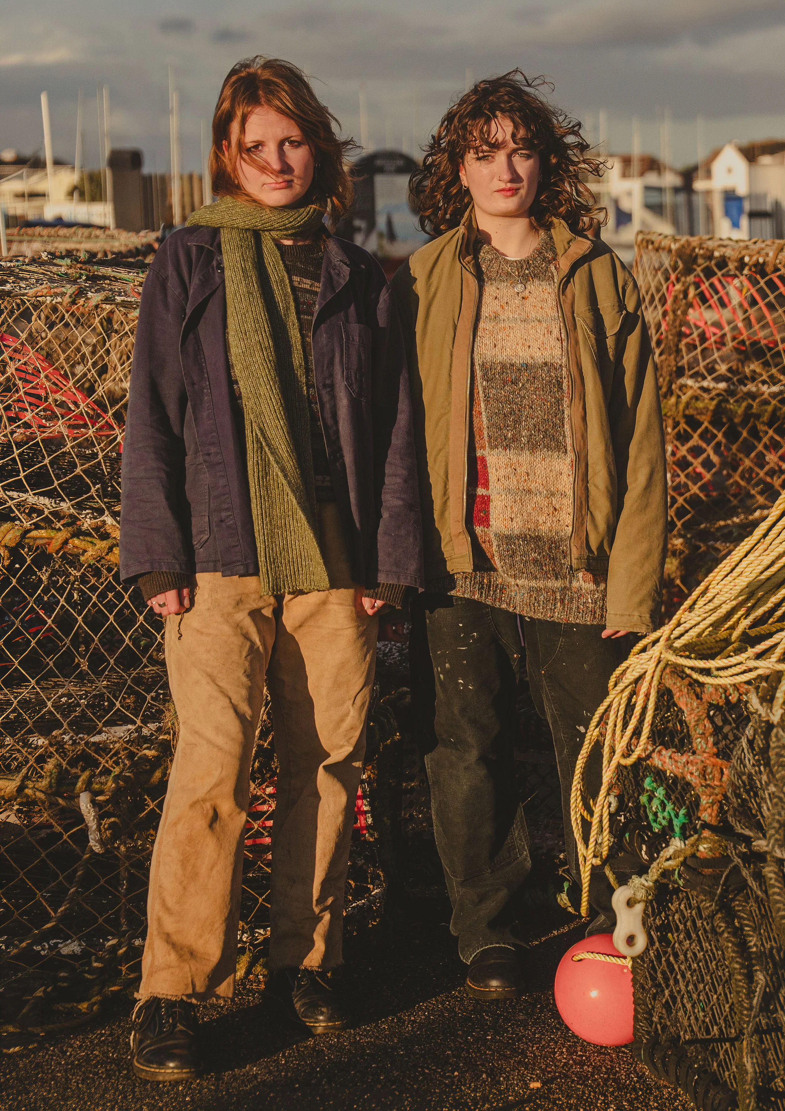 Two women standing outdoors near a fishing boat with a net and float in the foreground and a harbor with boats and houses in the background, during sunset.