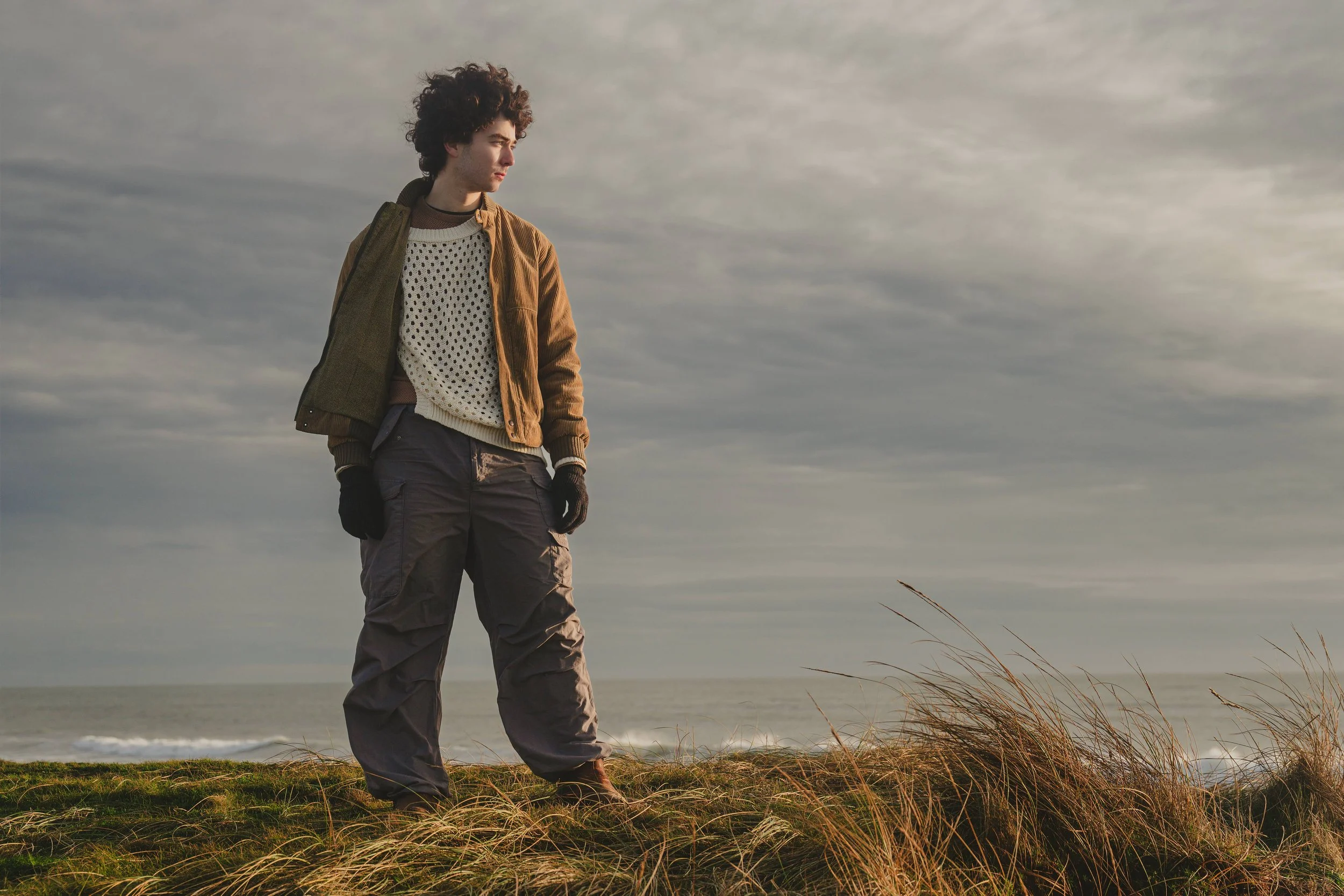 A young man standing outdoors near the ocean during cloudy weather, wearing a brown jacket, a white perforated sweater, dark cargo pants, and gloves, looking towards the sky.
