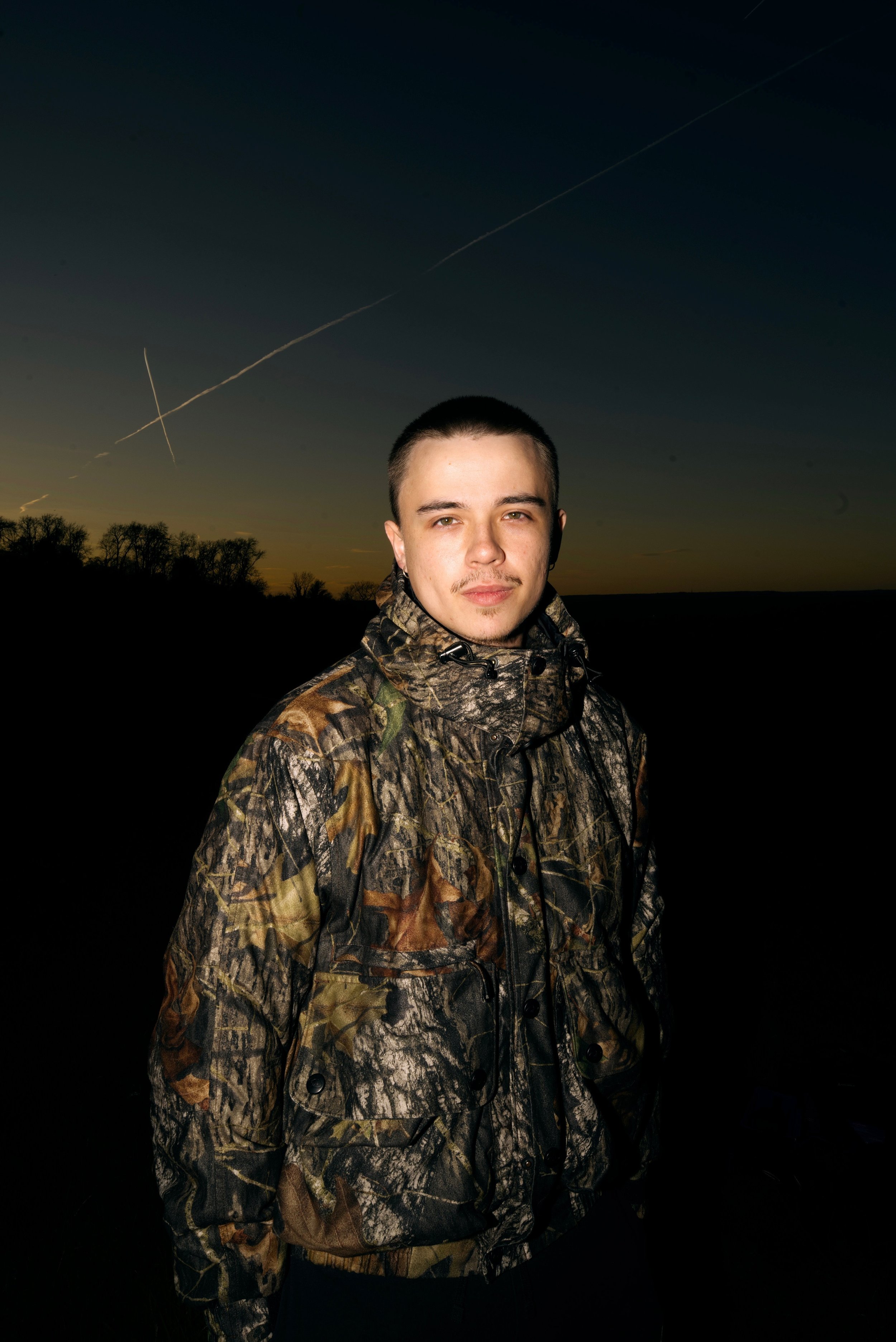 A young man with short dark hair and a slight mustache, wearing a camouflage jacket, standing outdoors during sunset or dusk, with trees and contrails in the darkening sky.