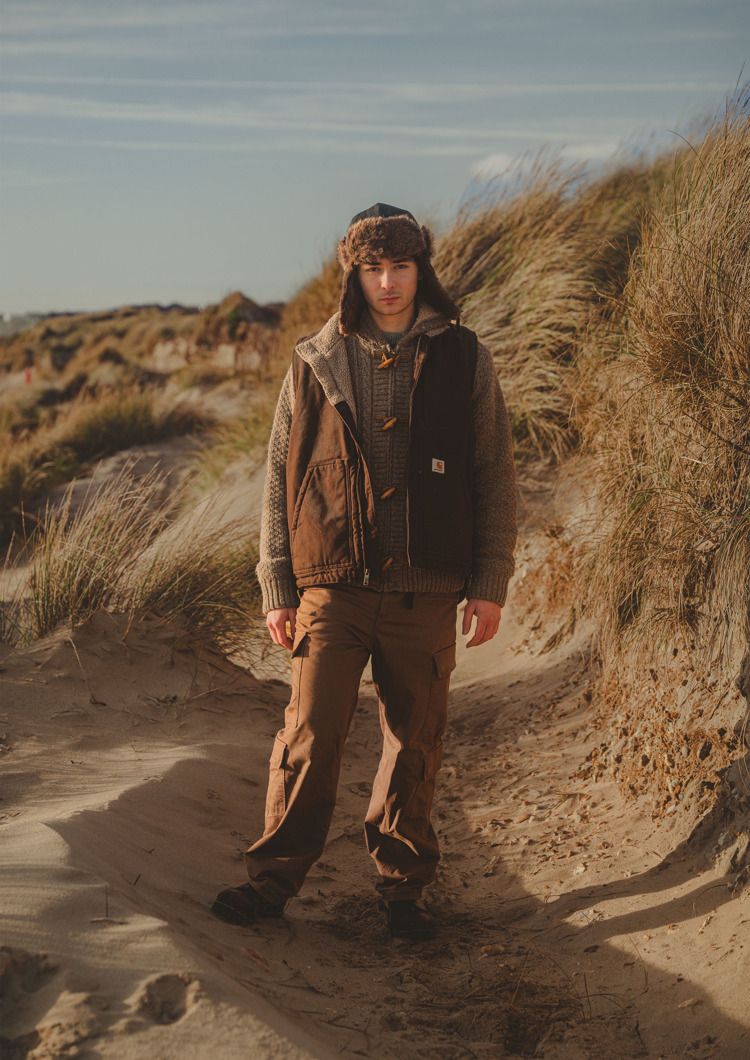 Young man standing on sandy beach with tall grass dunes behind him, dressed in outdoor winter clothing including a fur hat and brown pants.