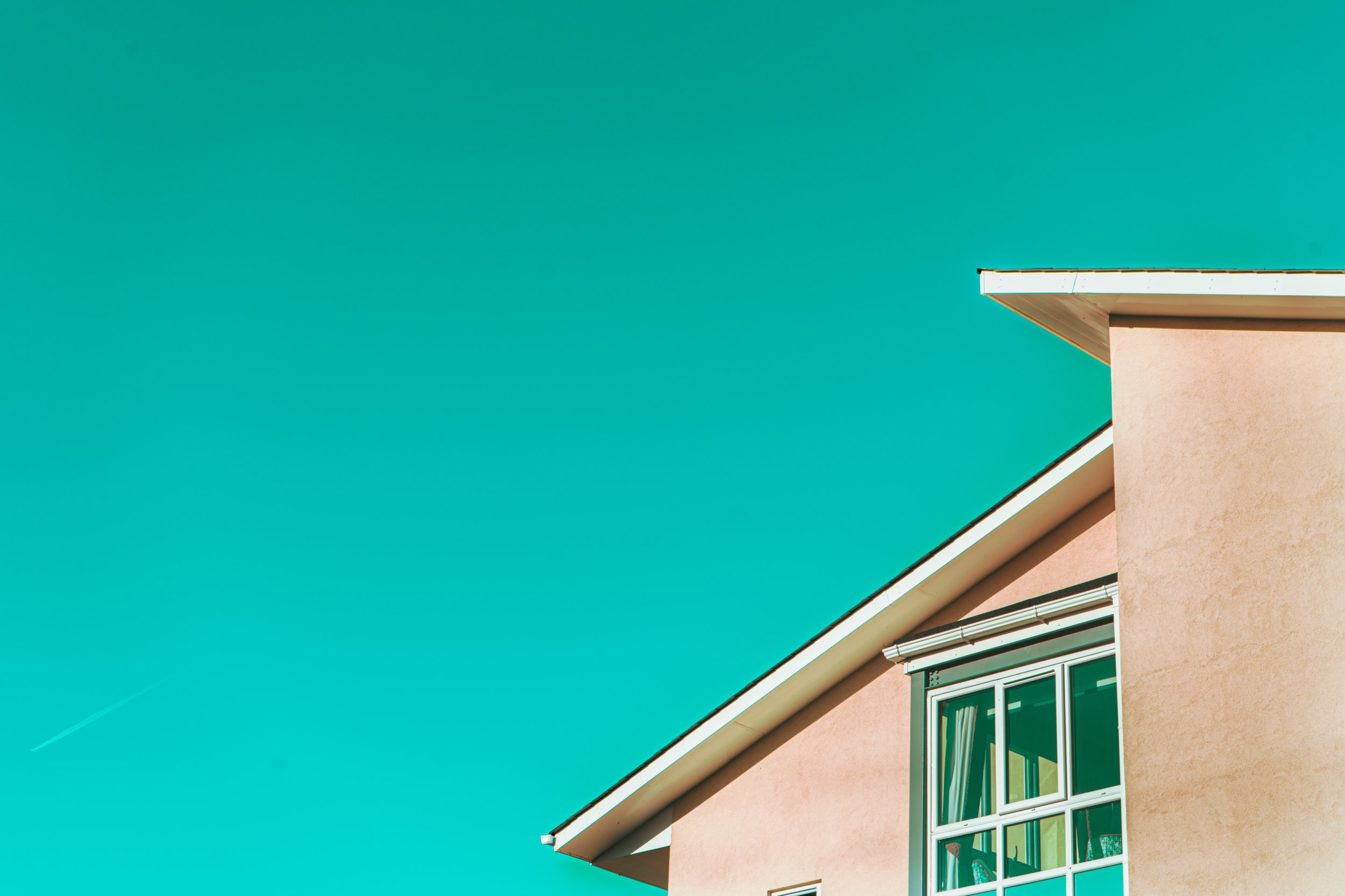 A building corner with a peach wall, window, and slanted roof against a clear turquoise sky.