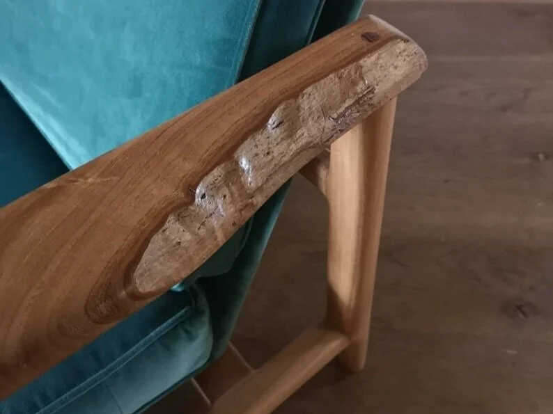 A wooden table with a natural, uneven edge sits next to a teal upholstered chair on a wooden floor.