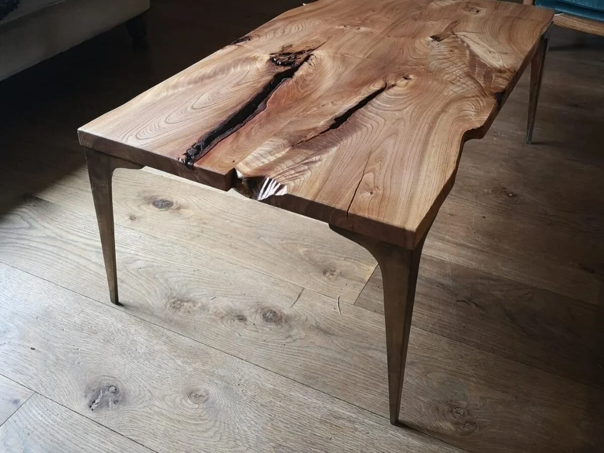 A wooden table with a natural, live edge design and visible grain patterns, supported by four slender legs, set on a wooden floor.