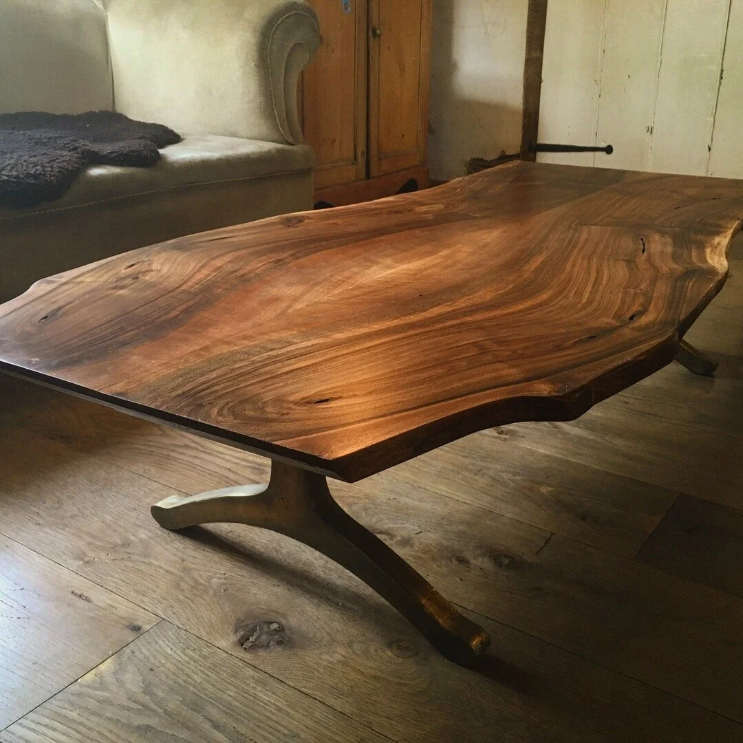 A rustic wooden table with natural wood grain, curved edges, and a sturdy base in a cozy room with wooden floors and furniture.