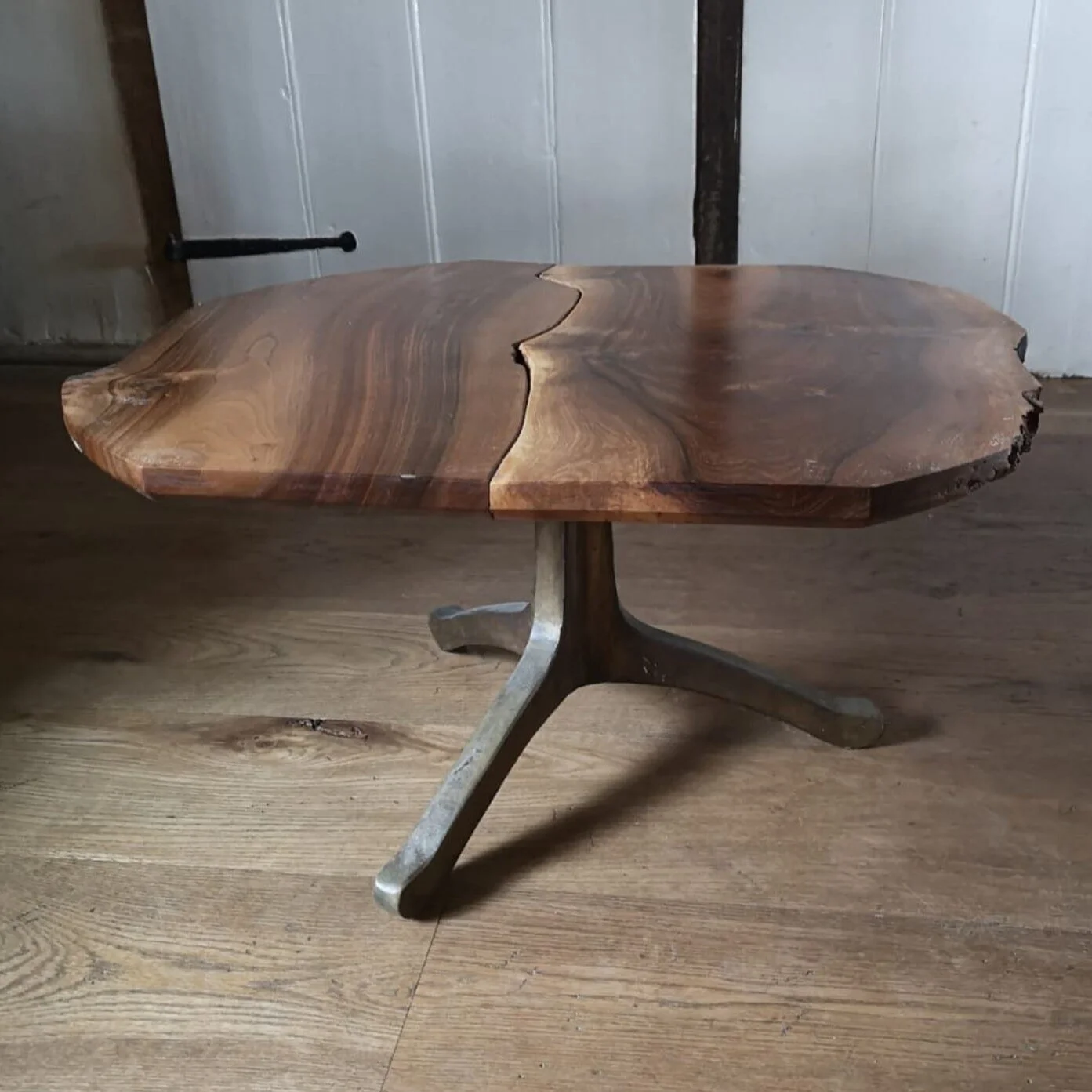 A rustic wooden table with a split down the middle, supported by a metal leg with three feet, placed on a hardwood floor next to a white wall and a black latch.