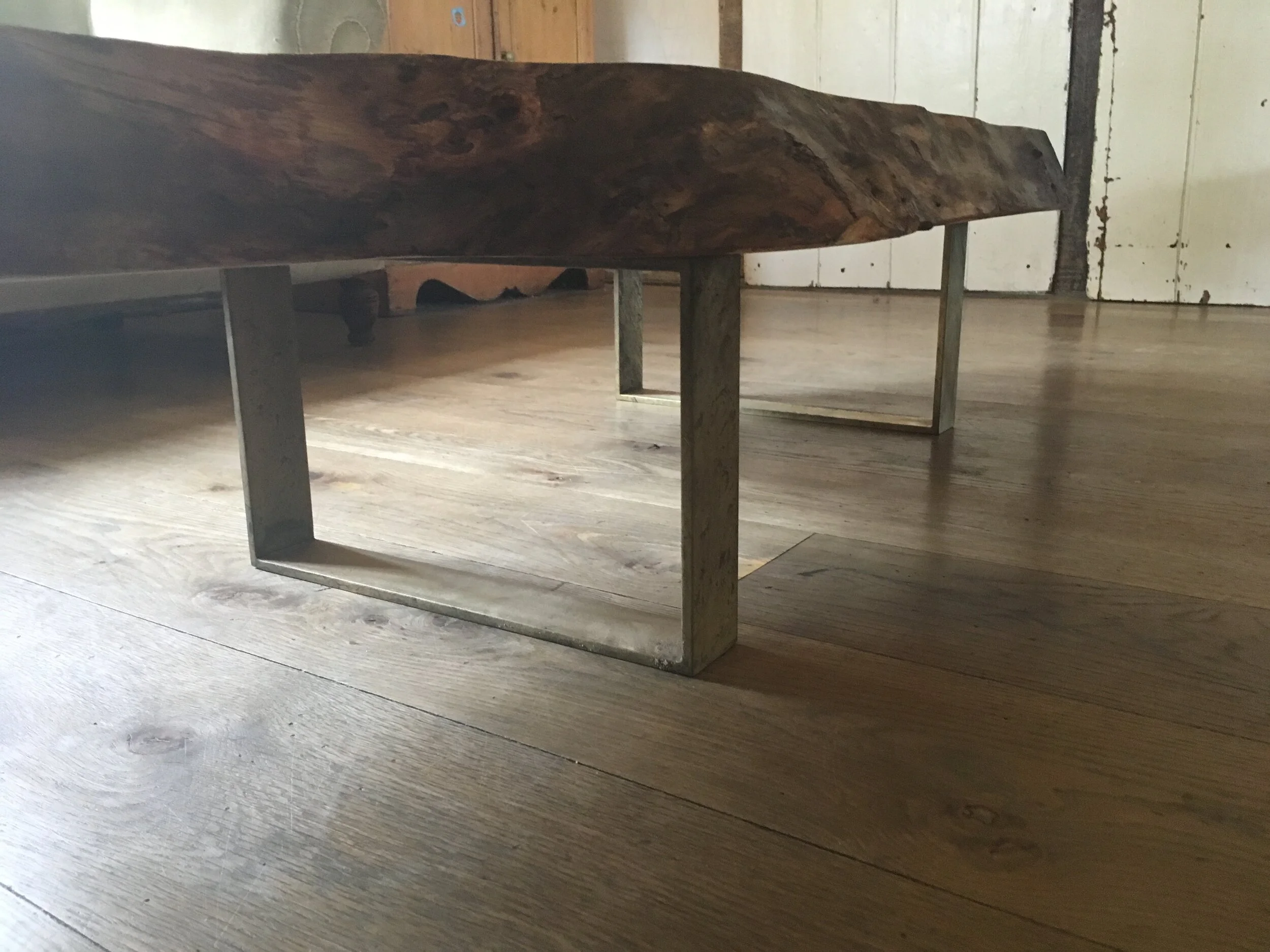 Close-up of a wooden and metal coffee table with uneven natural wood tabletop and geometric metal legs, placed on a wooden floor indoors.