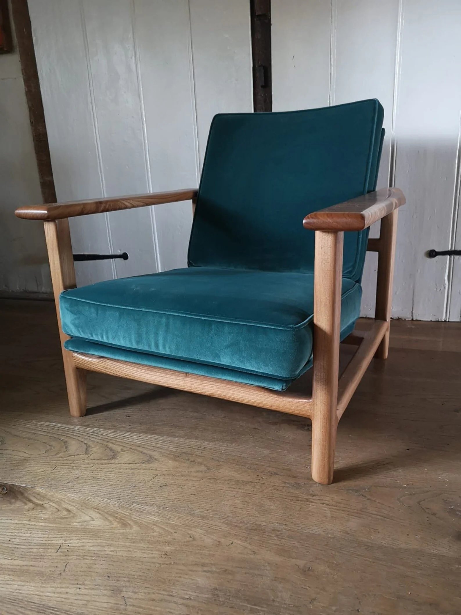 A vintage armchair with teal velvet cushions and wooden frame and armrests on a wooden floor with white paneled wall background.