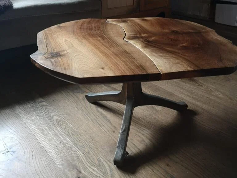 A round wooden table with a natural, live edge top and a four-legged metal base, placed on a hardwood floor.
