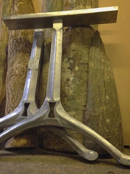 A metallic chair or stool with a uniquely shaped frame, placed in front of a pair of large, textured wooden logs.