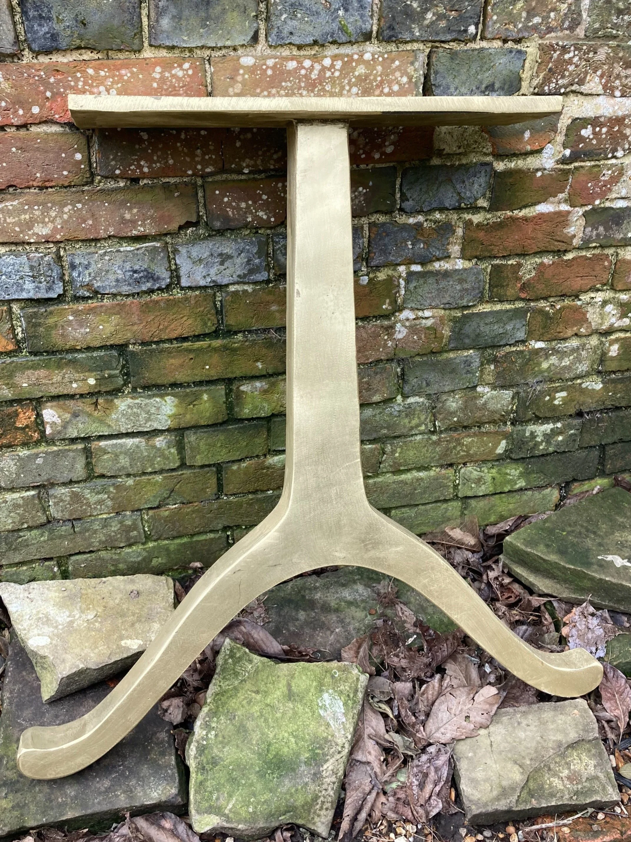 A gold-colored metal tree stand for a Christmas tree placed on uneven stones and fallen leaves in front of a brick wall.