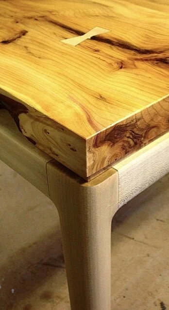 Close-up of a wooden table corner showing a natural wood tabletop and a beige upholstered chair leg underneath.