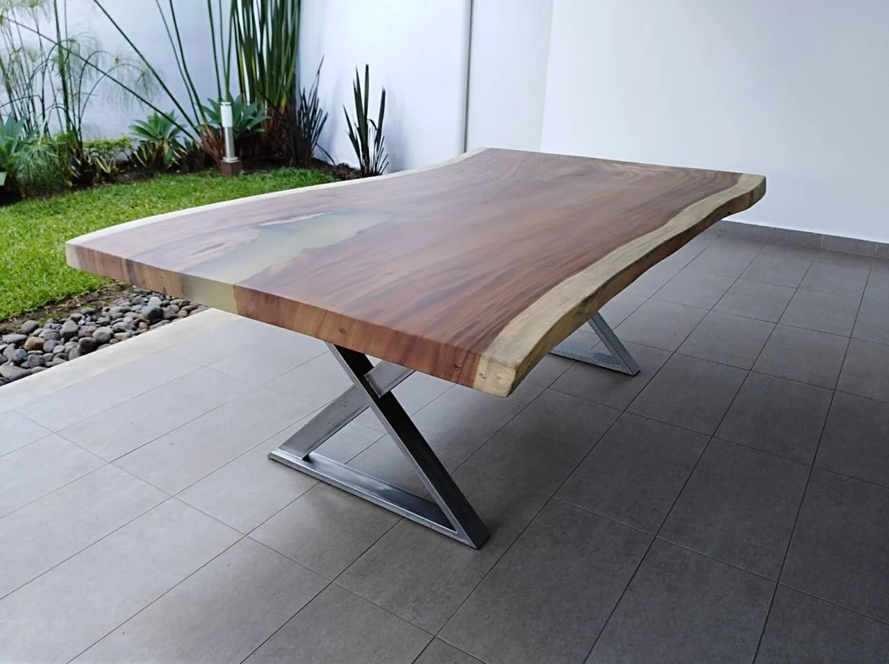 A modern wooden table with a live edge on a gray tiled patio, with a garden and white wall in the background.