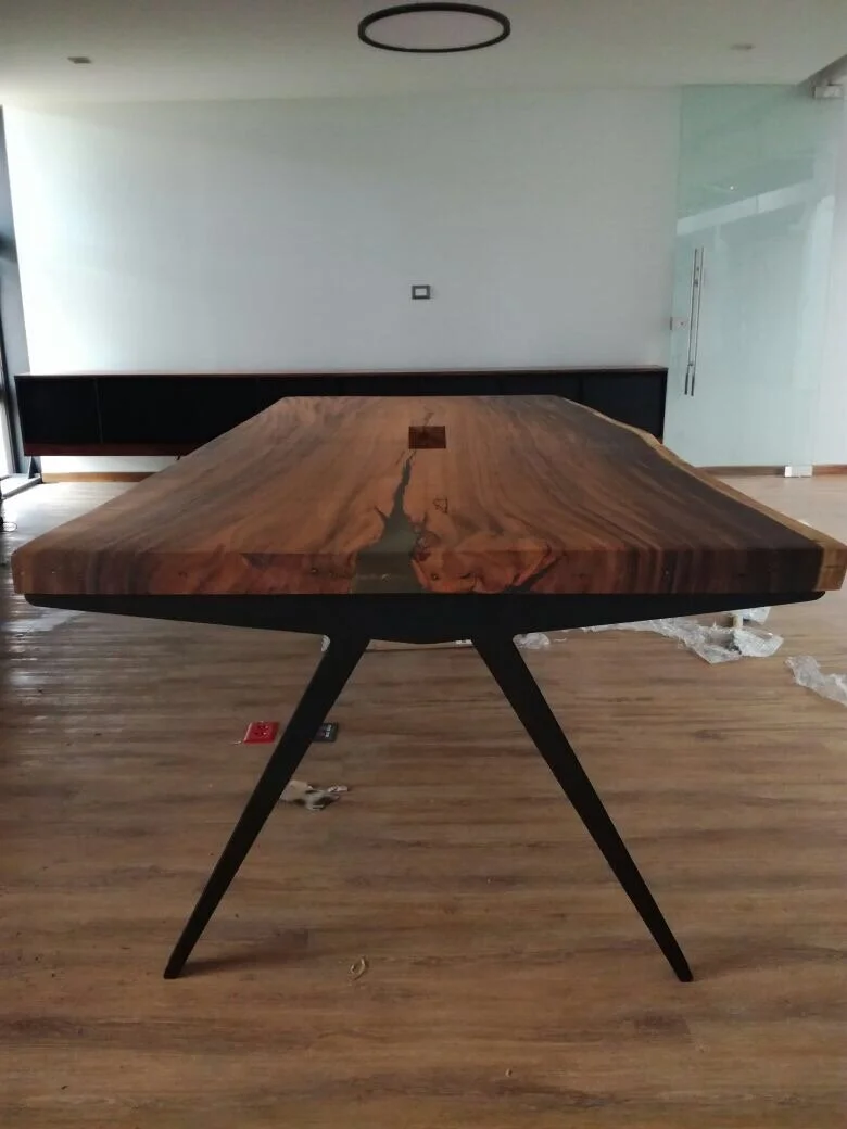 A large, modern wooden dining table with a natural edge, supported by black angled legs, in a spacious room with wooden flooring and minimal decor.