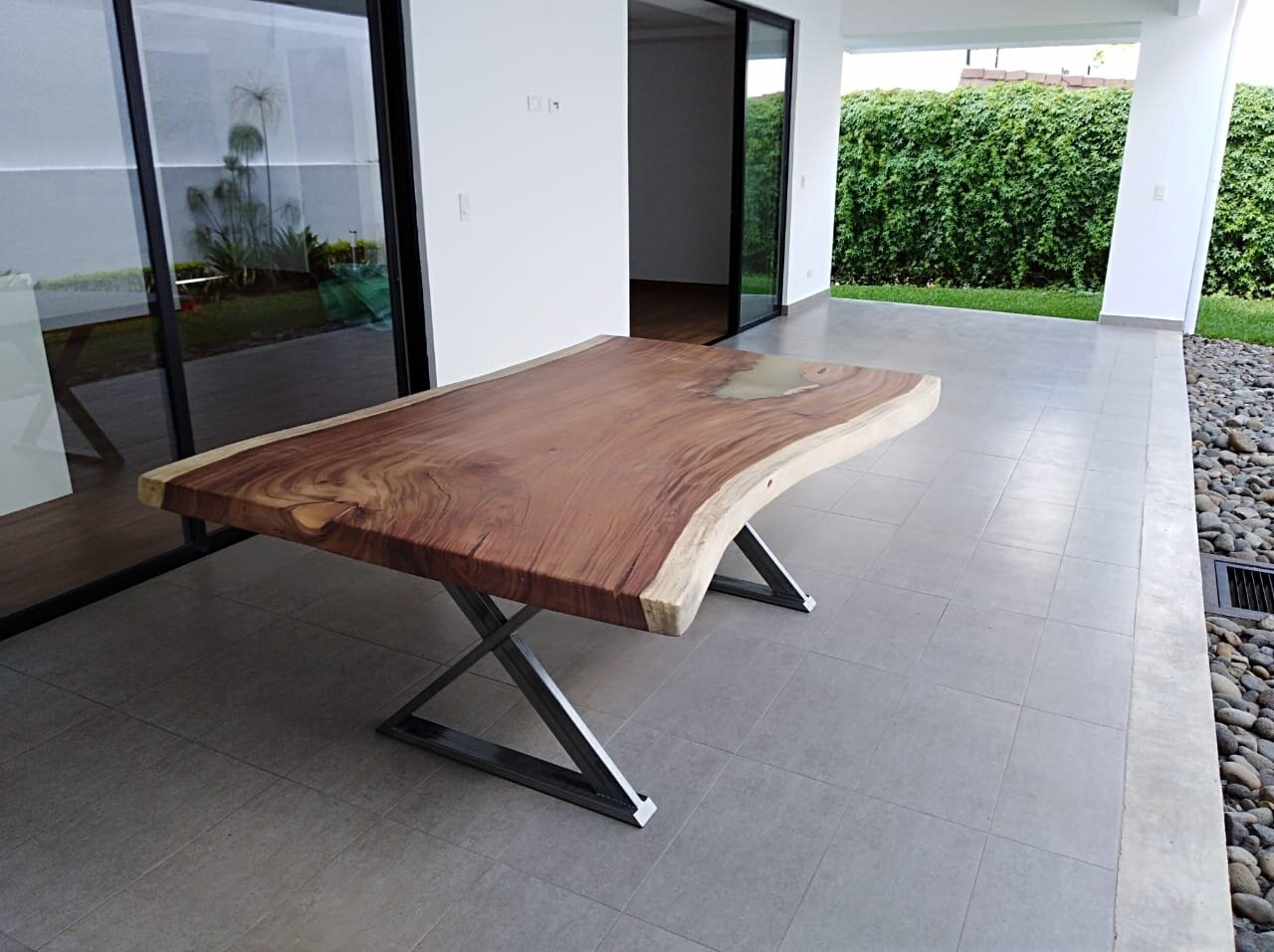 Modern covered outdoor patio with a live edge wooden table and lush green hedge in the background.