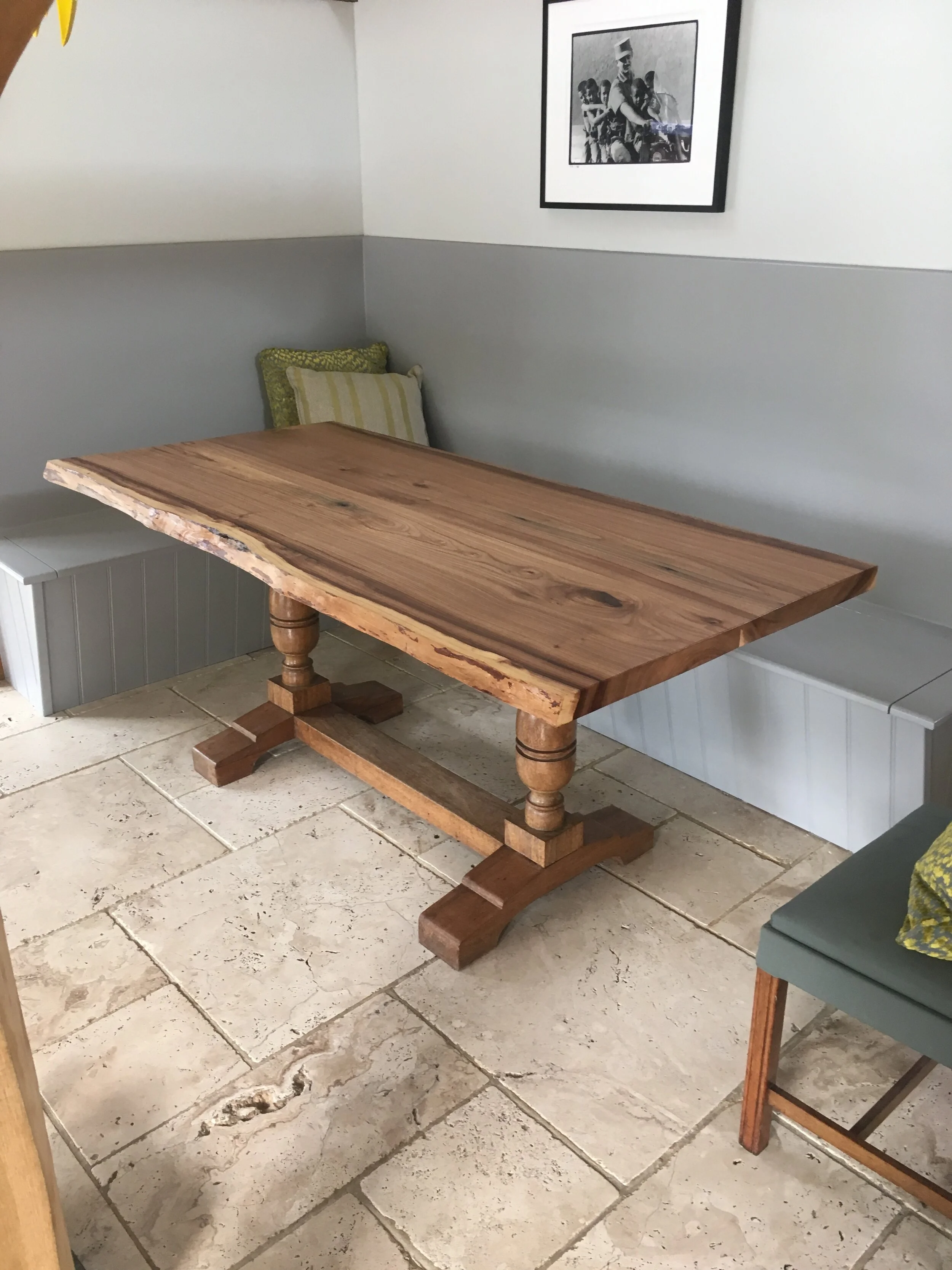 A rustic wooden dining table with turned legs and a natural edge, situated in a corner with a bench and a chair, in a room with tiled flooring and a black-and-white framed photograph on the wall.