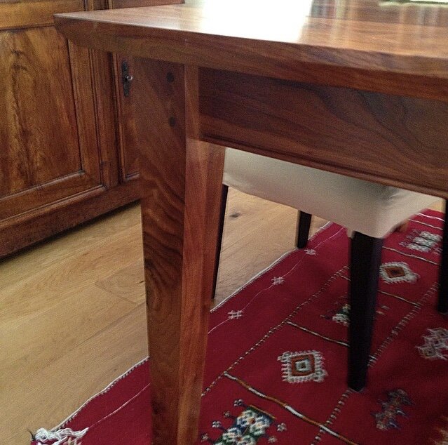 Close-up of a wooden table leg with a smooth finish, showing the edge of the tabletop, a chair with a white cushion, and a red patterned rug on a hardwood floor.