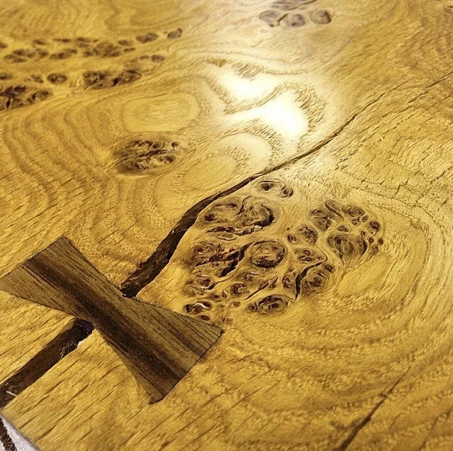 Close-up of a wooden table with natural knots and grain patterns, and a small wooden wedge inserted into a crack.