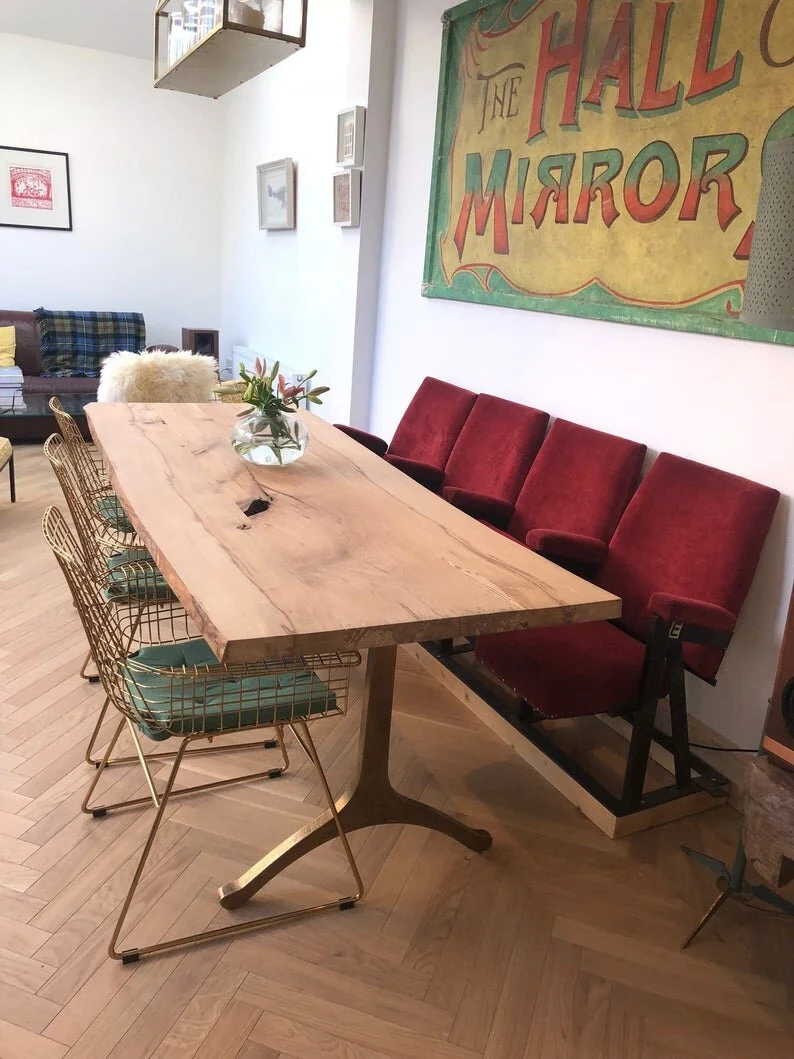 A dining area with a large wooden table, six green wireframe chairs with cushions, and a red vintage theater-style bench against the wall. There is a vase with flowers on the table and colorful artwork on the walls.